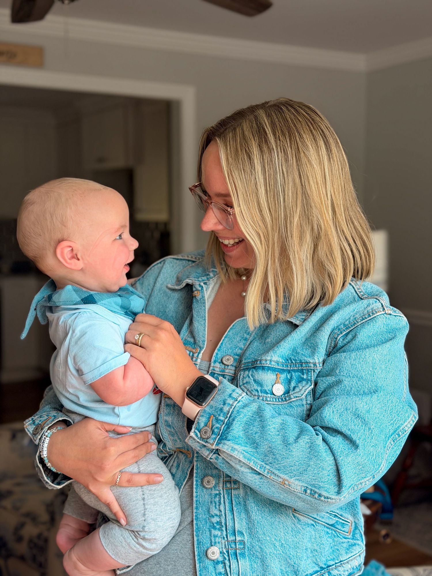 Smiling blonde woman in a denim jacket holding and interacting with a happy baby indoors.