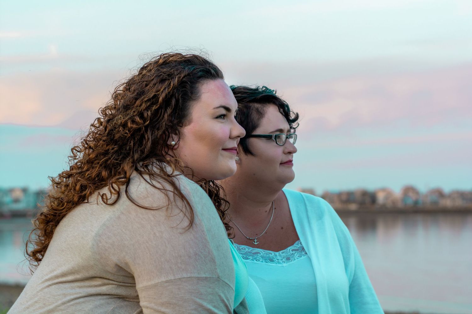 Two women gazing at a pastel sunset by the waterfront, one with curly hair in a beige cardigan, the other in a mint top.