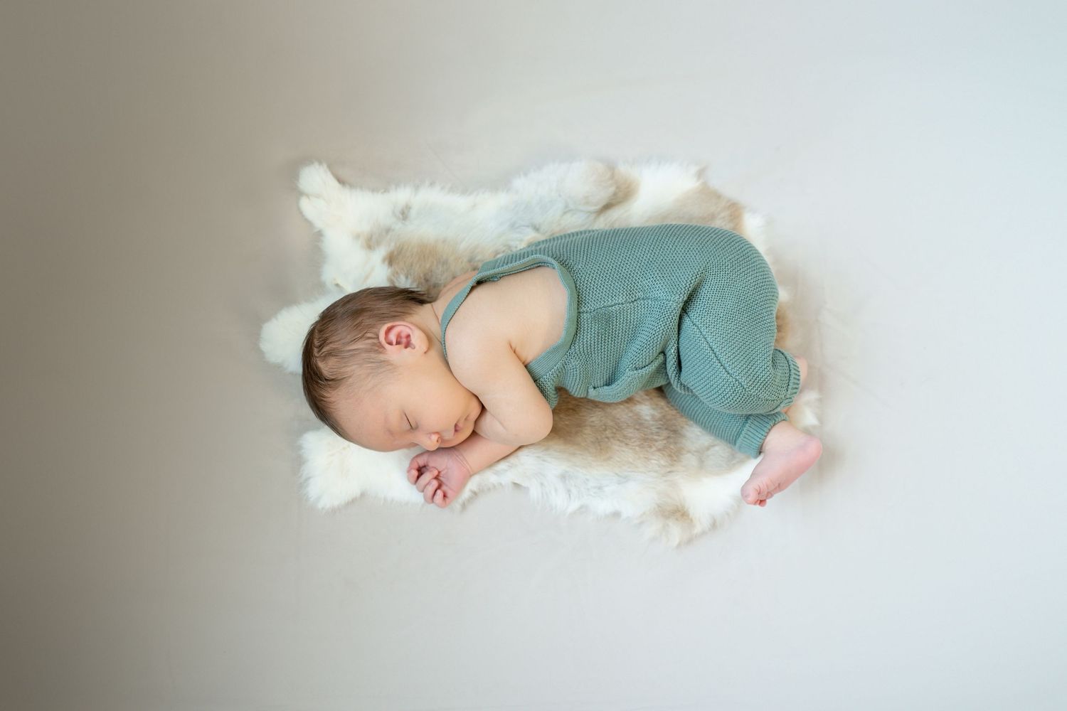 Peaceful newborn baby sleeping on fluffy white fur in sage green romper, photographed from above.