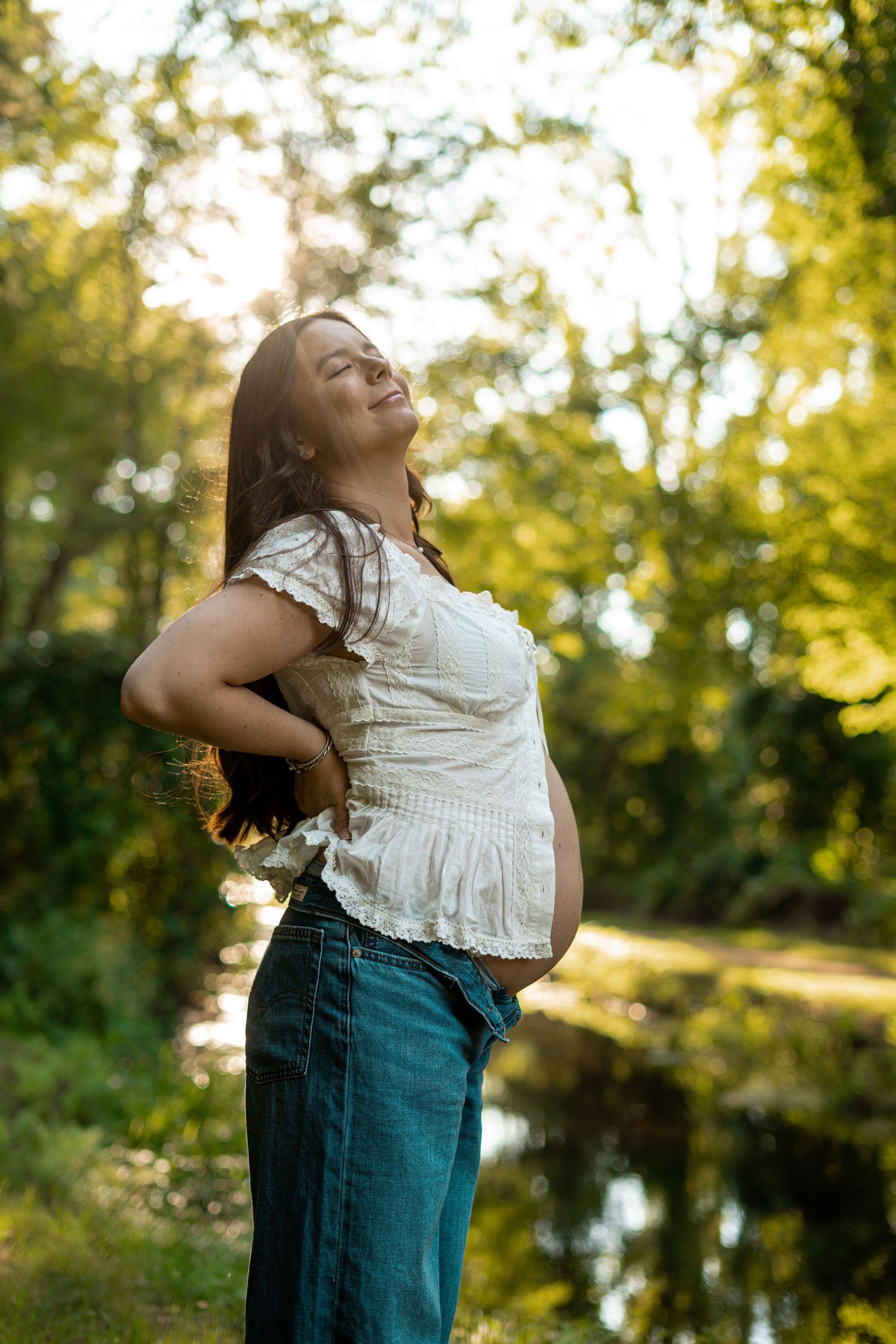 Radiant pregnant woman in white top and jeans poses with eyes closed in golden sunlit outdoor setting.