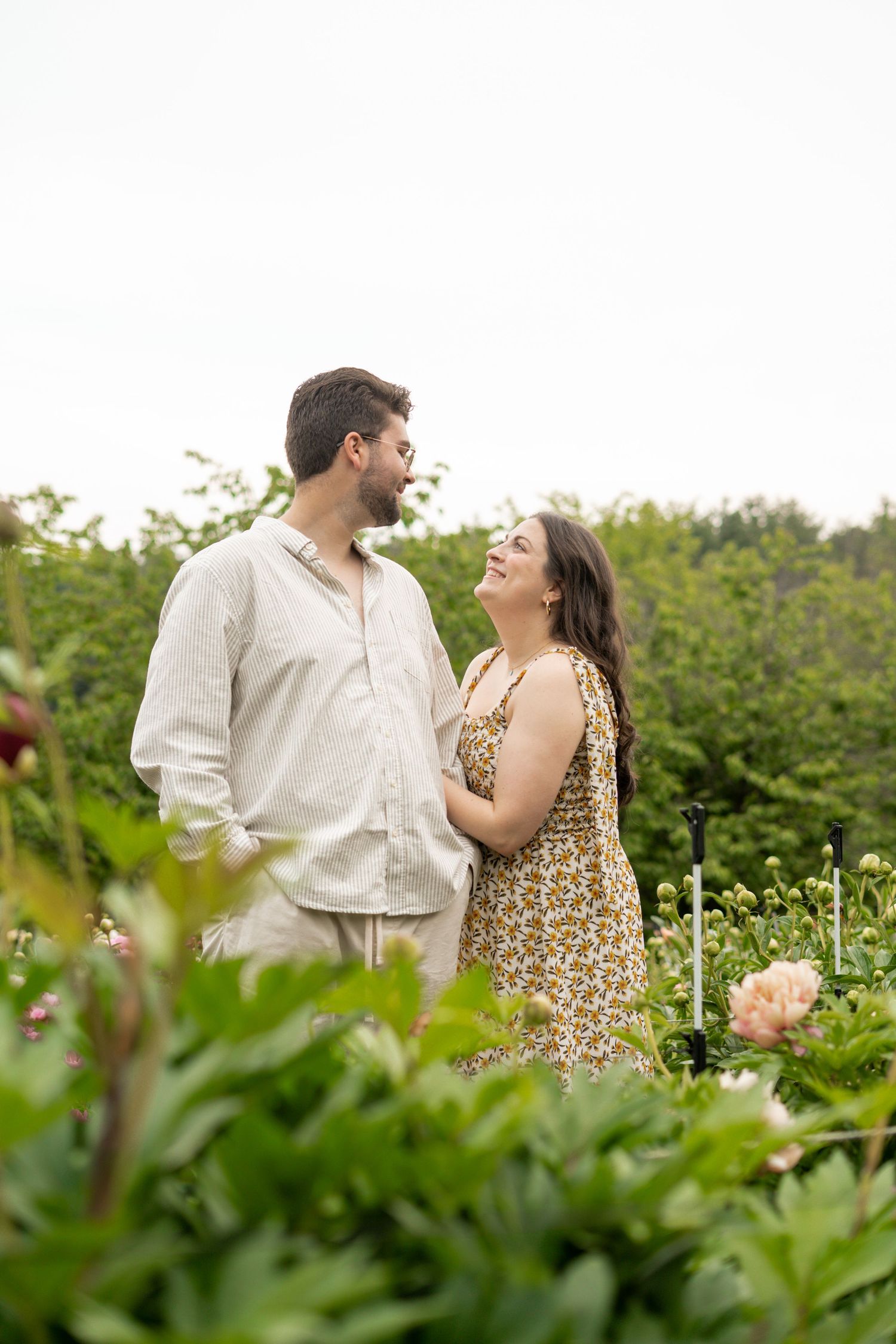 Couple gazing at each other lovingly while standing among lush green peony garden plants.