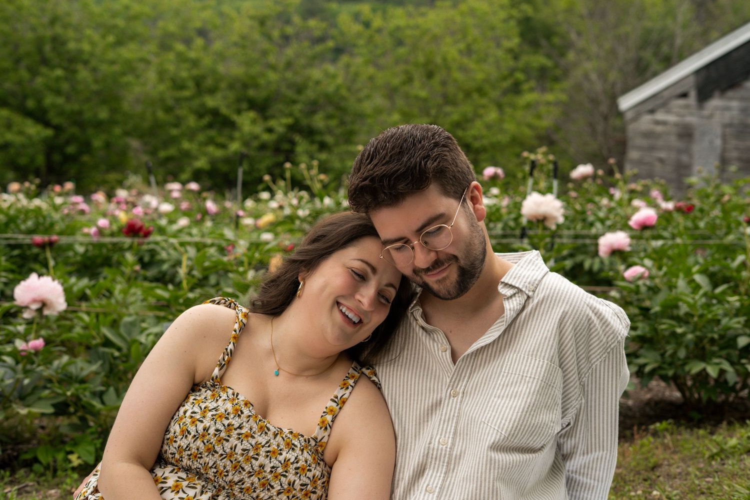 Happy couple leaning together and smiling in front of blooming pink peony garden.