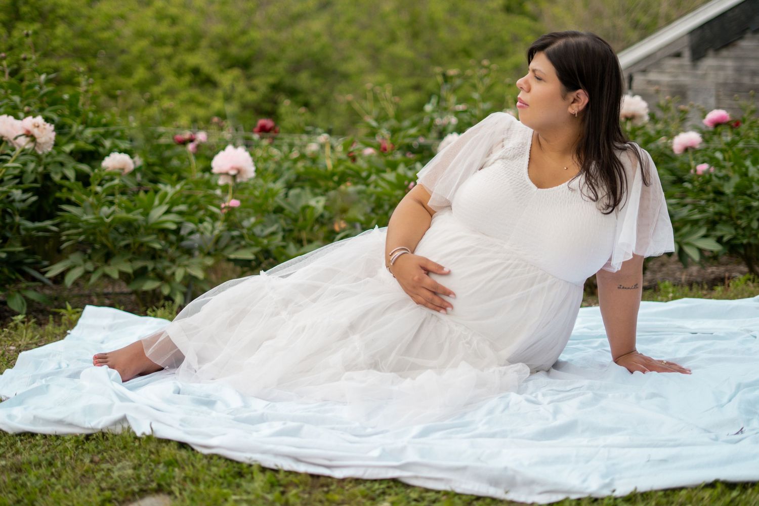 Pregnant woman in white dress sitting on blanket in garden with pink peonies blooming in background.