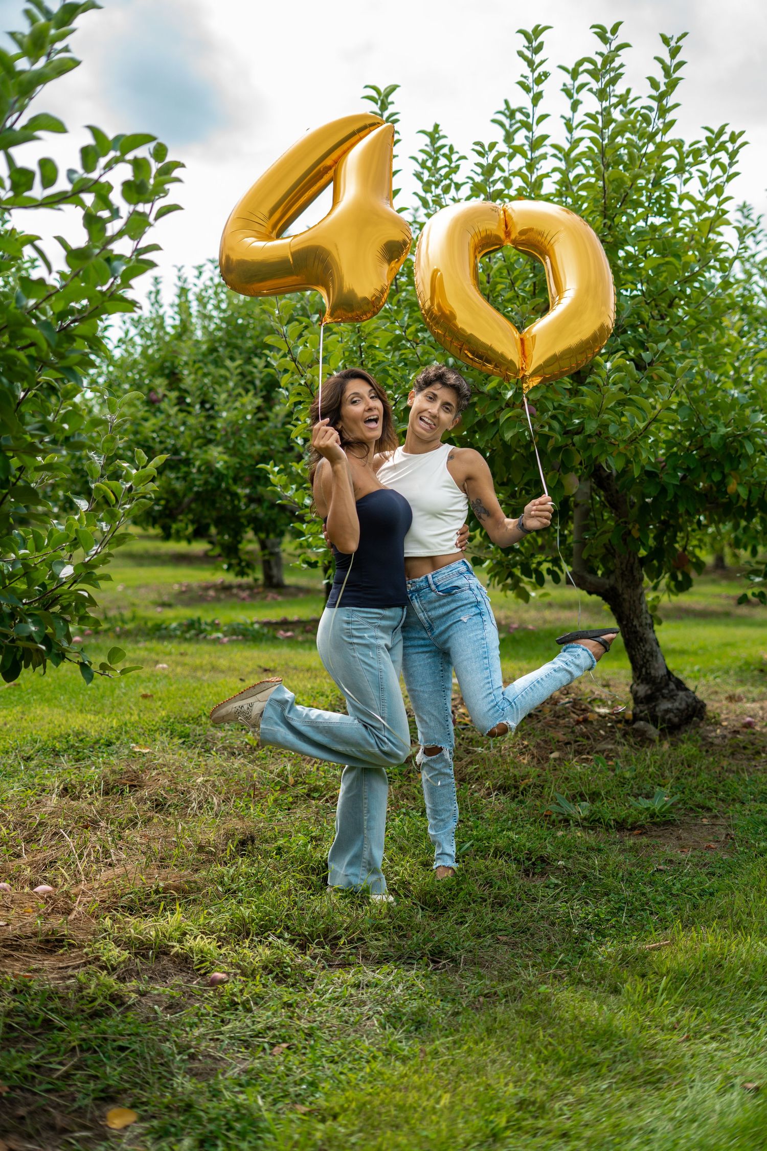 Two joyful women holding gold number 40 balloons celebrating a birthday in an orchard.