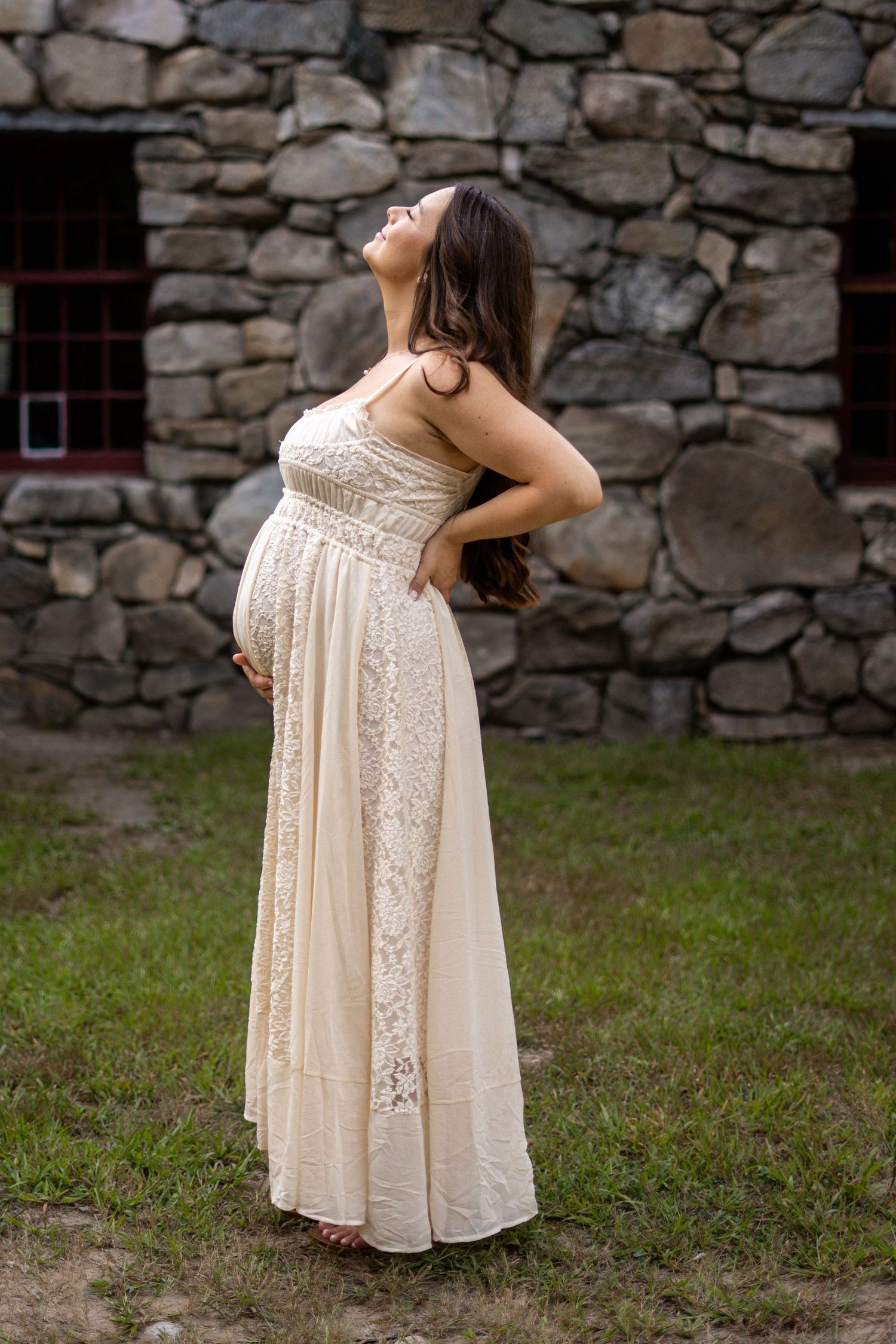 A pregnant woman in a cream lace maternity gown poses gracefully against a rustic stone wall outdoors.