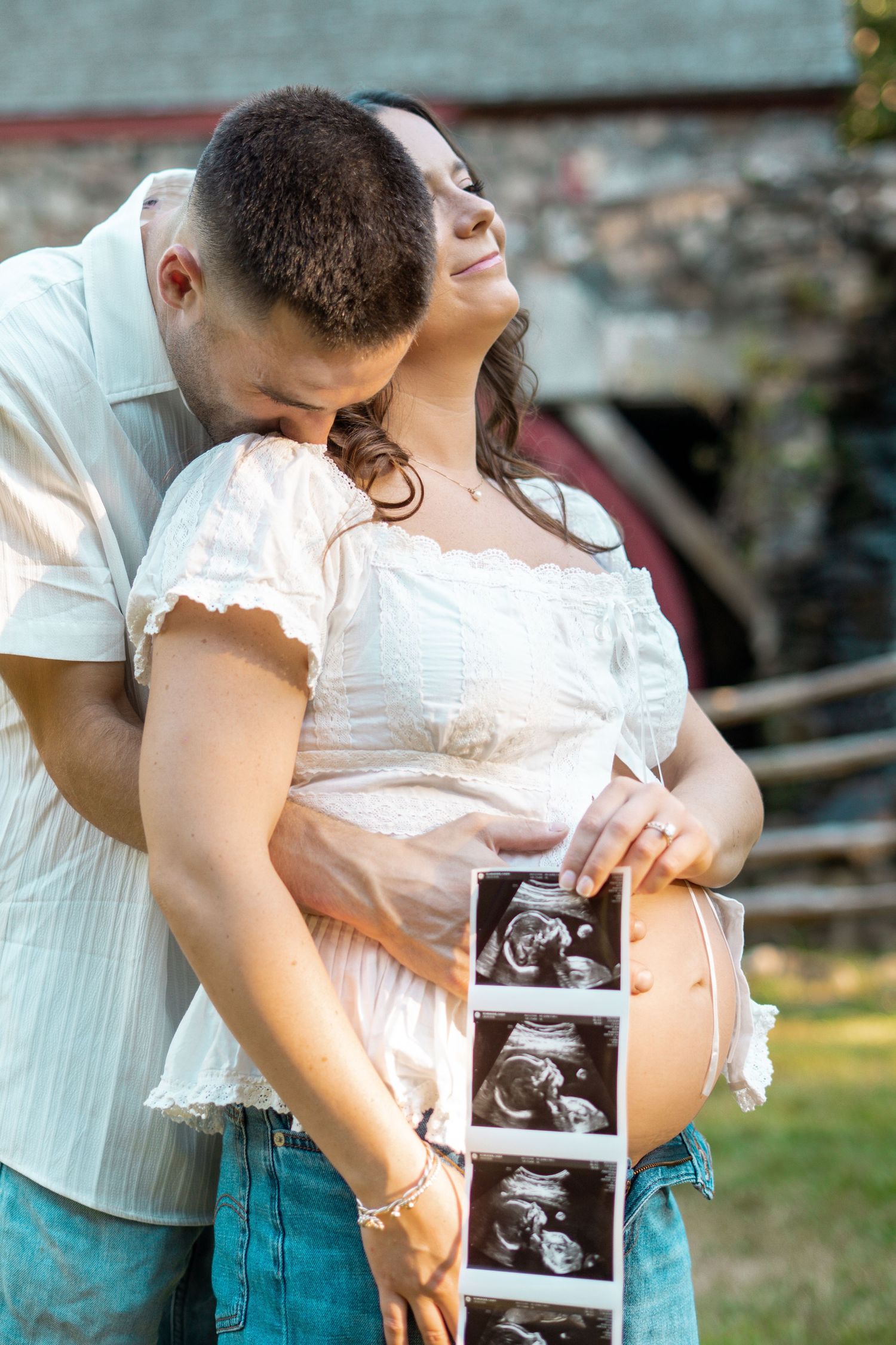 Couple shares emotional maternity moment holding ultrasound photos near rustic stone watermill outdoors.