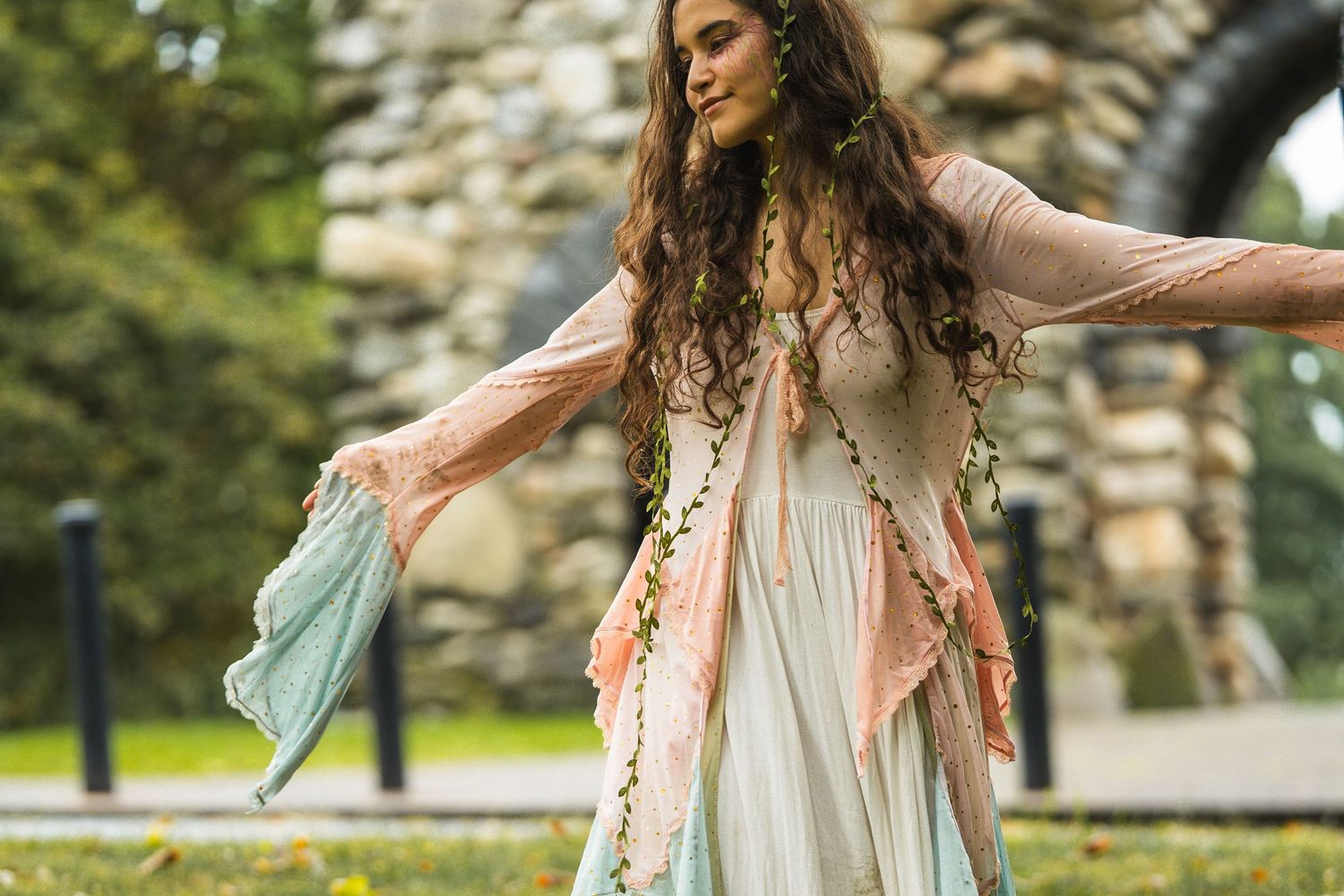 A woman in a flowing bohemian fairy costume with pastel layers and long curly hair dances outdoors in a park.