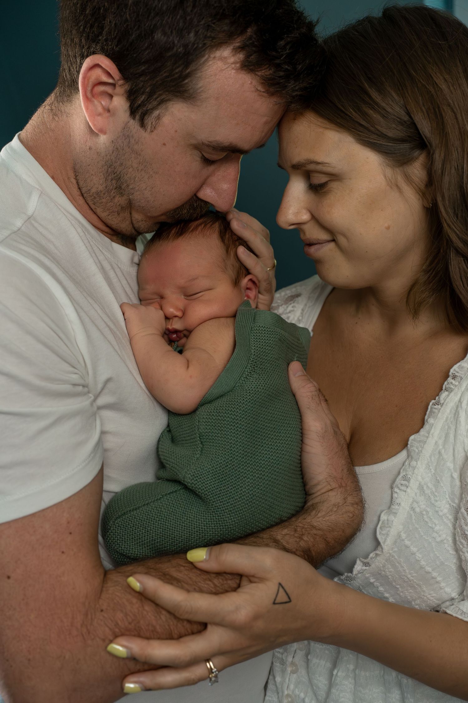 New parents holding their sleeping newborn wrapped in green against a teal background indoors.