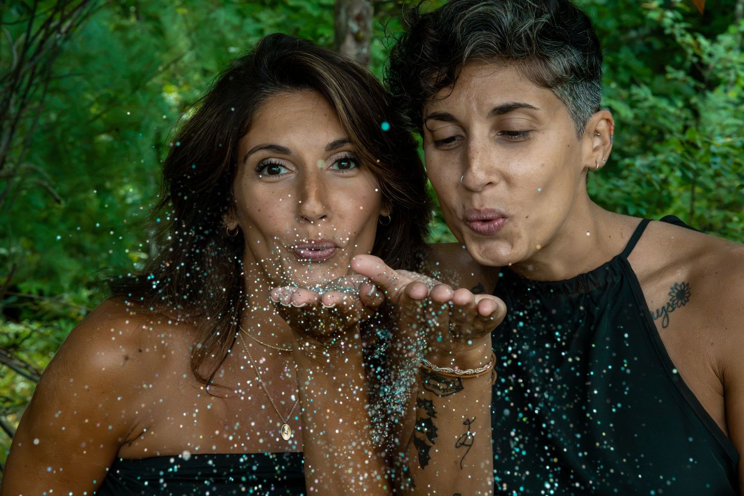 Two women blowing glitter from their hands outdoors surrounded by lush green trees.