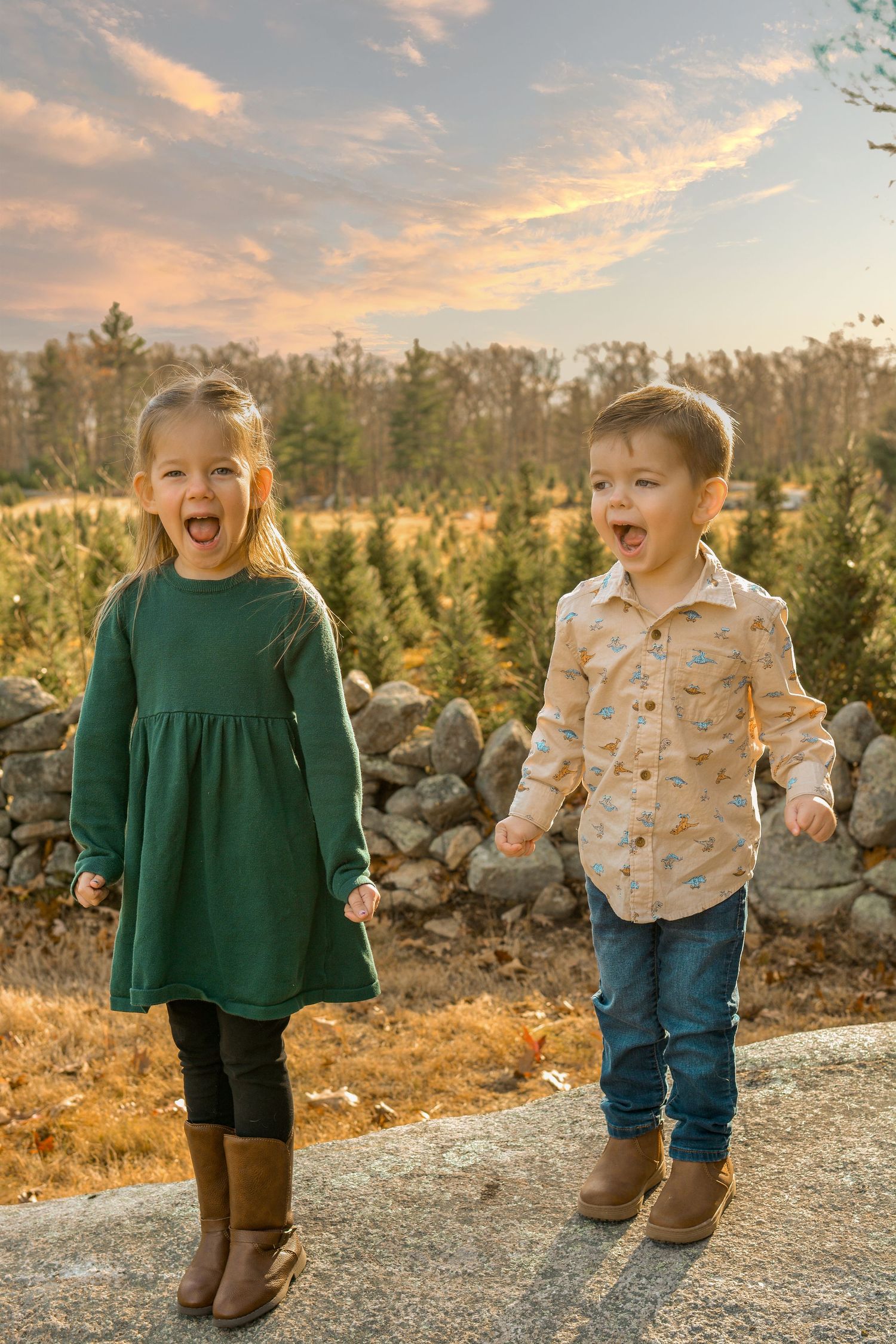 Two young children laughing outdoors at sunset near a Christmas tree farm, girl in green dress and boy in printed shirt.