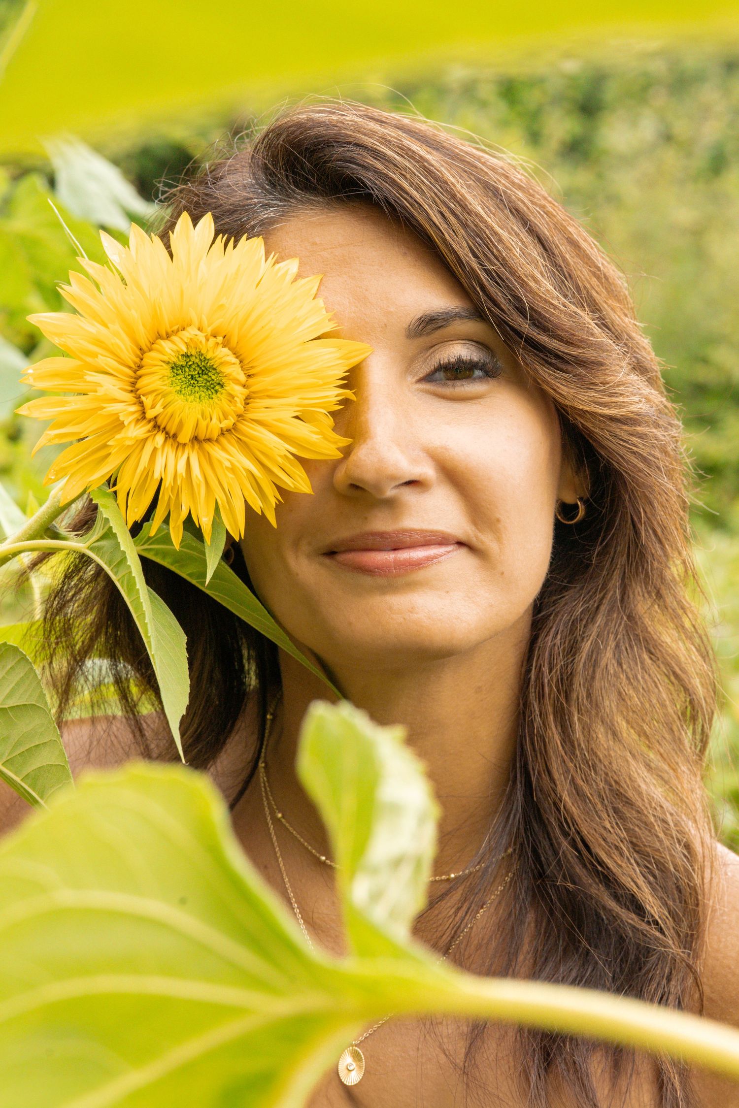 Beautiful woman holding a yellow sunflower over one eye in a sunny garden setting.