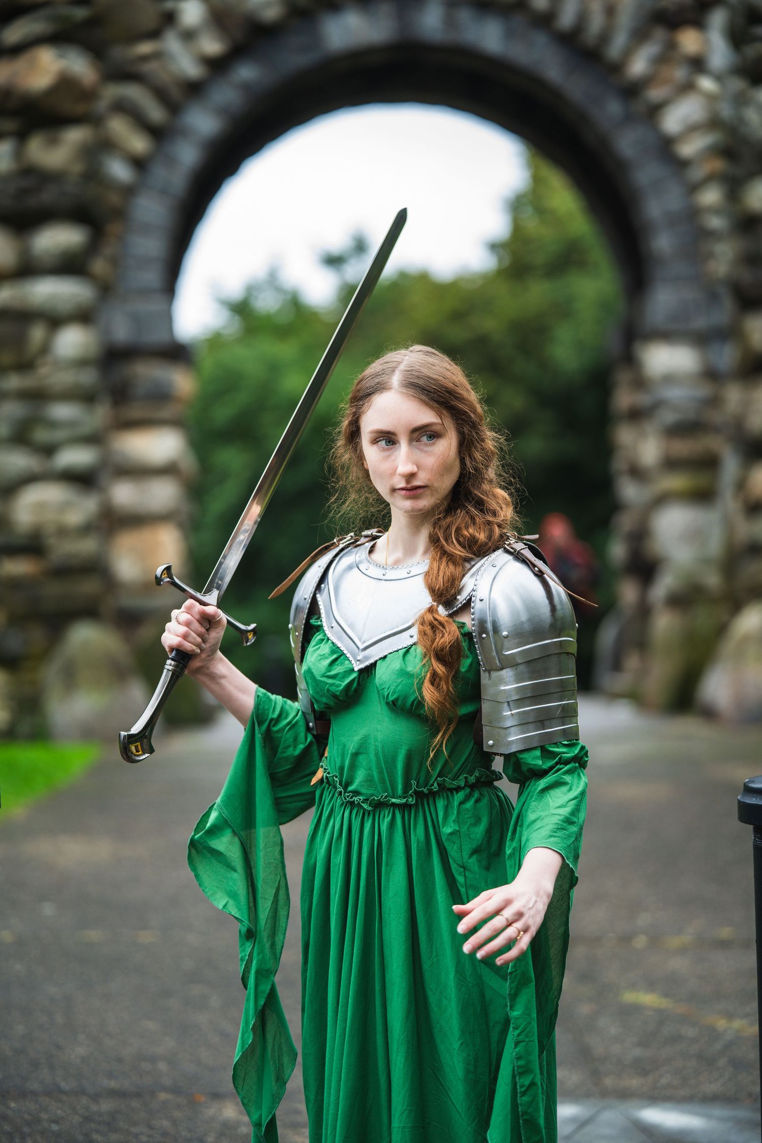 A woman in a green medieval dress and armor holds a sword before a stone archway.