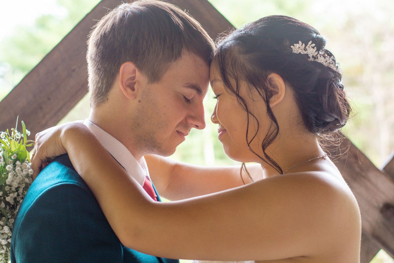 Bride and groom touching foreheads in a tender moment beneath a wooden arch at their wedding ceremony.