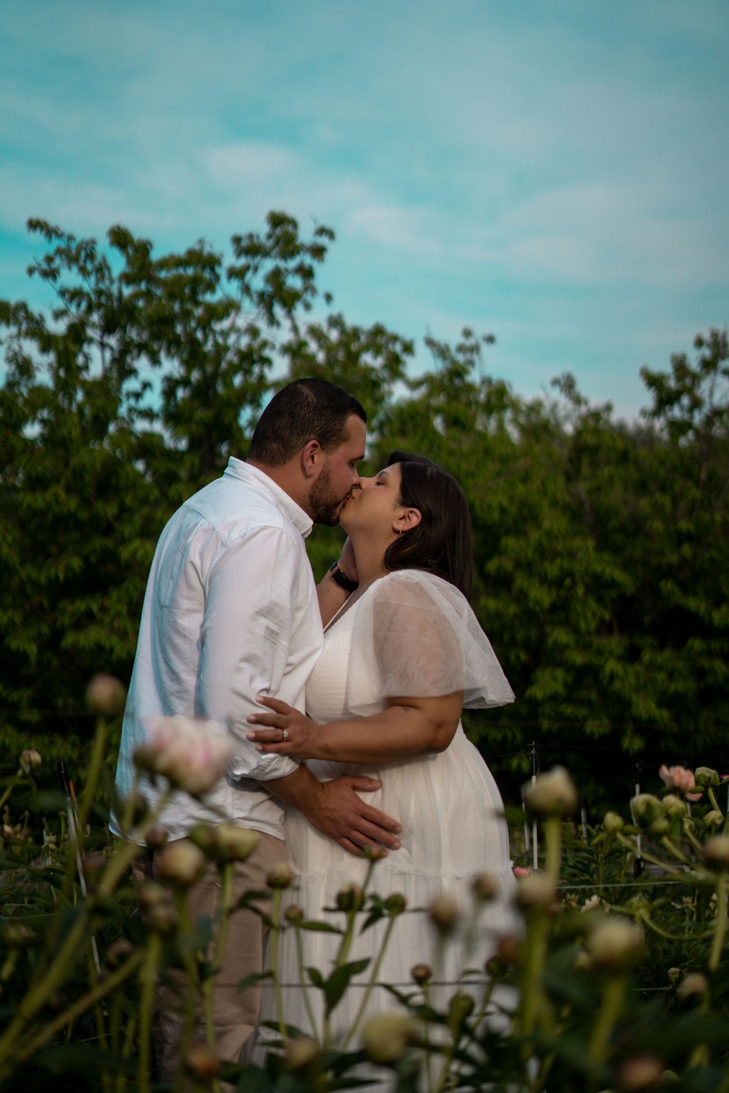 Couple sharing a kiss in flower field, pregnant woman in white dress held tenderly.