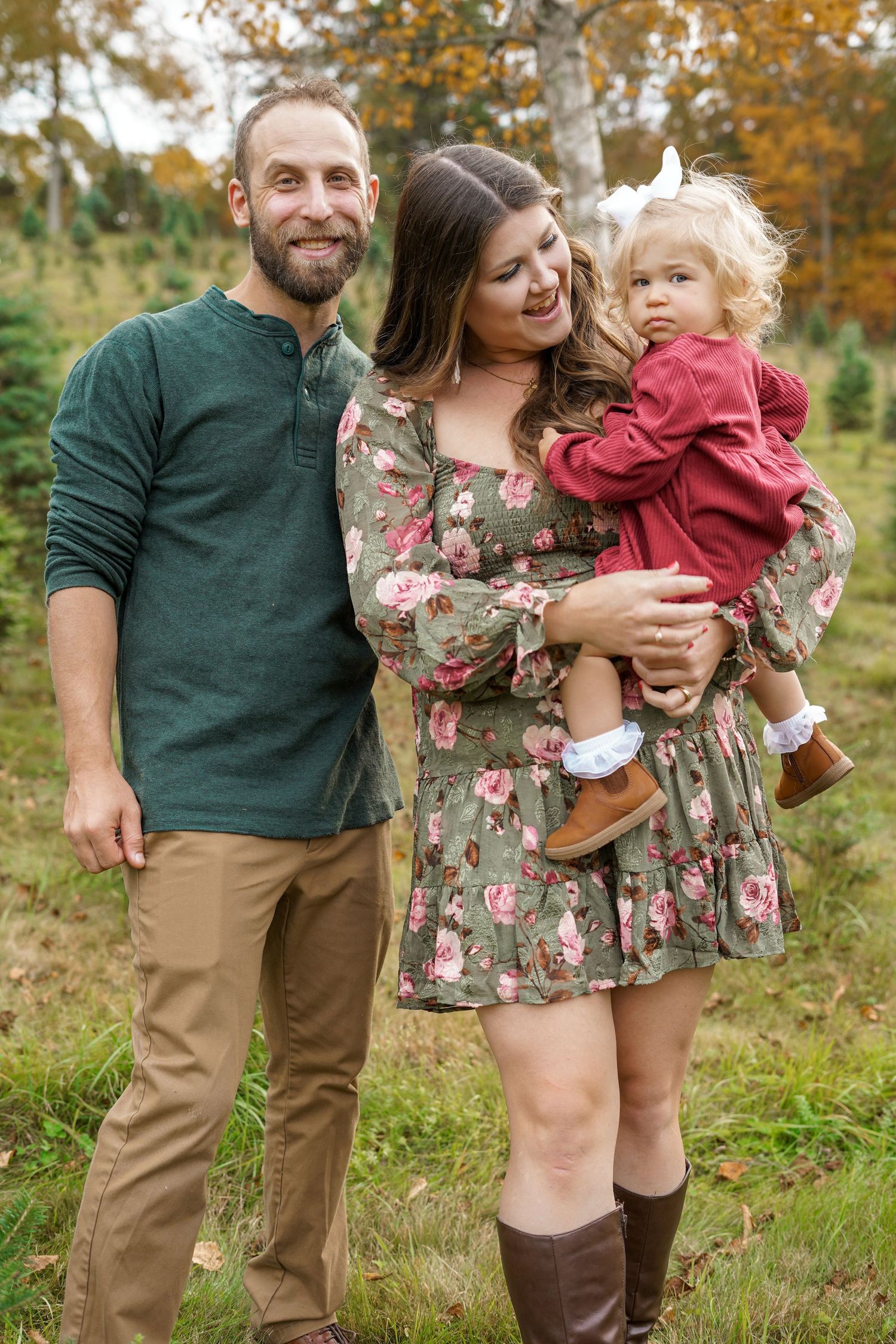 Family of three posing at Christmas tree farm in autumn, mother holding toddler in red dress.