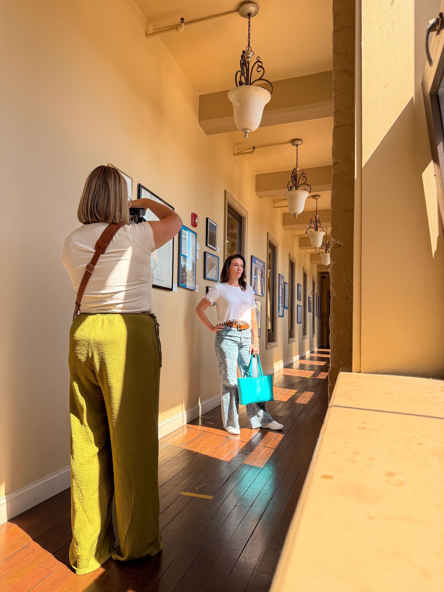 Blonde woman in olive pants photographing a brunette woman posing in a sunlit hallway gallery.