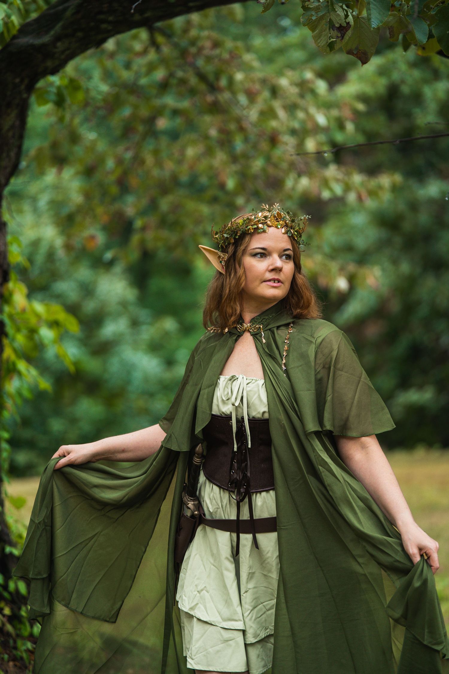 Woman dressed as a forest elf in green cloak, floral crown, and pointed ears, posing outdoors in a woodland setting.