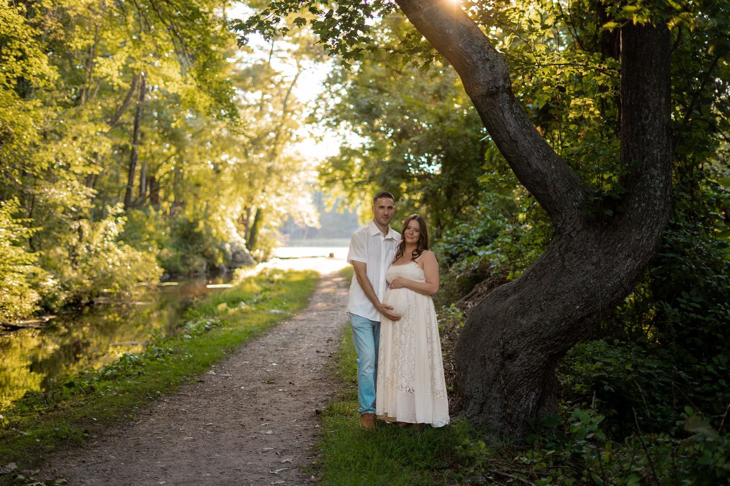 Expectant couple standing together on a sunlit forest path near a calm river, woman in white lace dress.