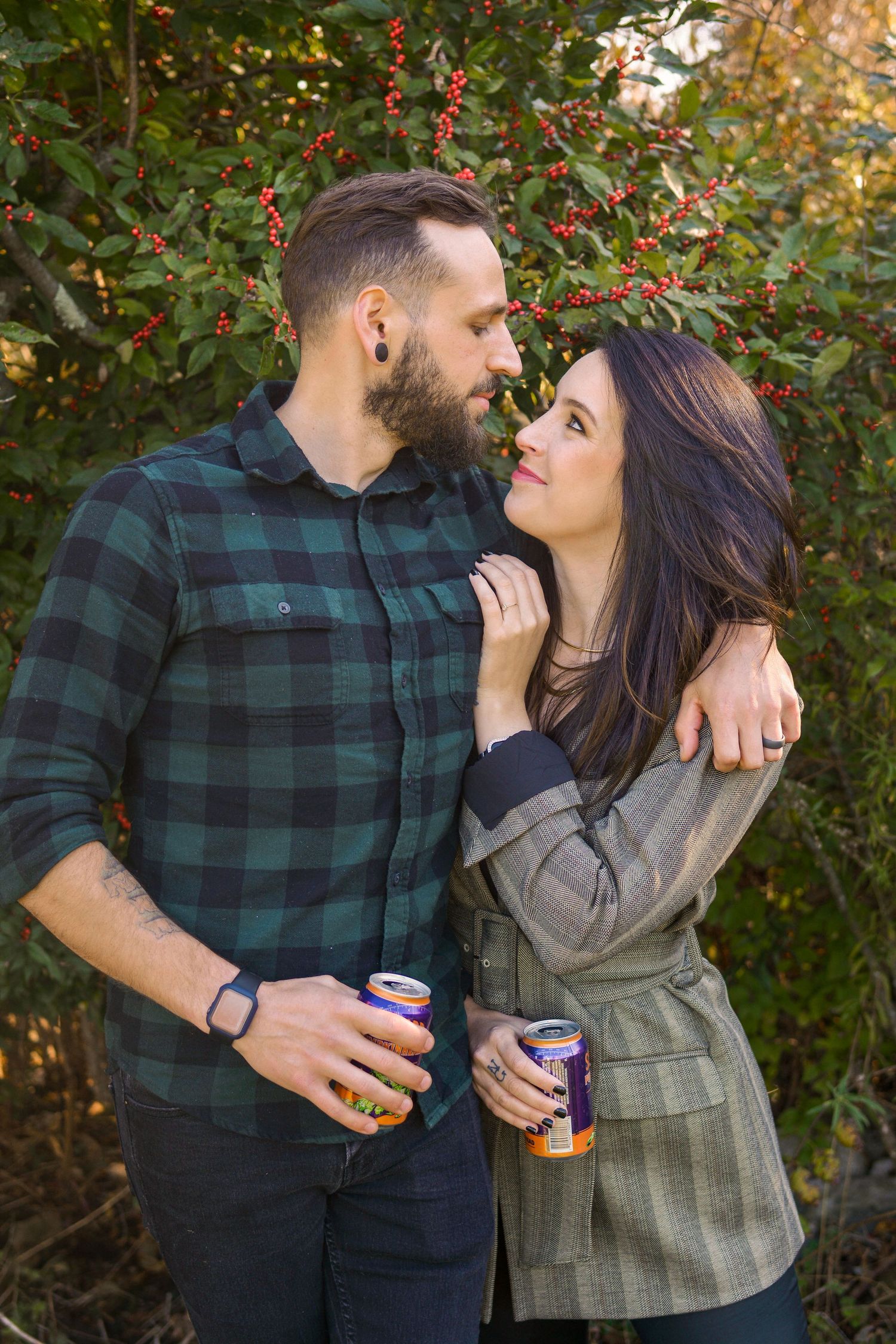 Romantic couple embracing and holding canned drinks near a red berry bush outdoors.
