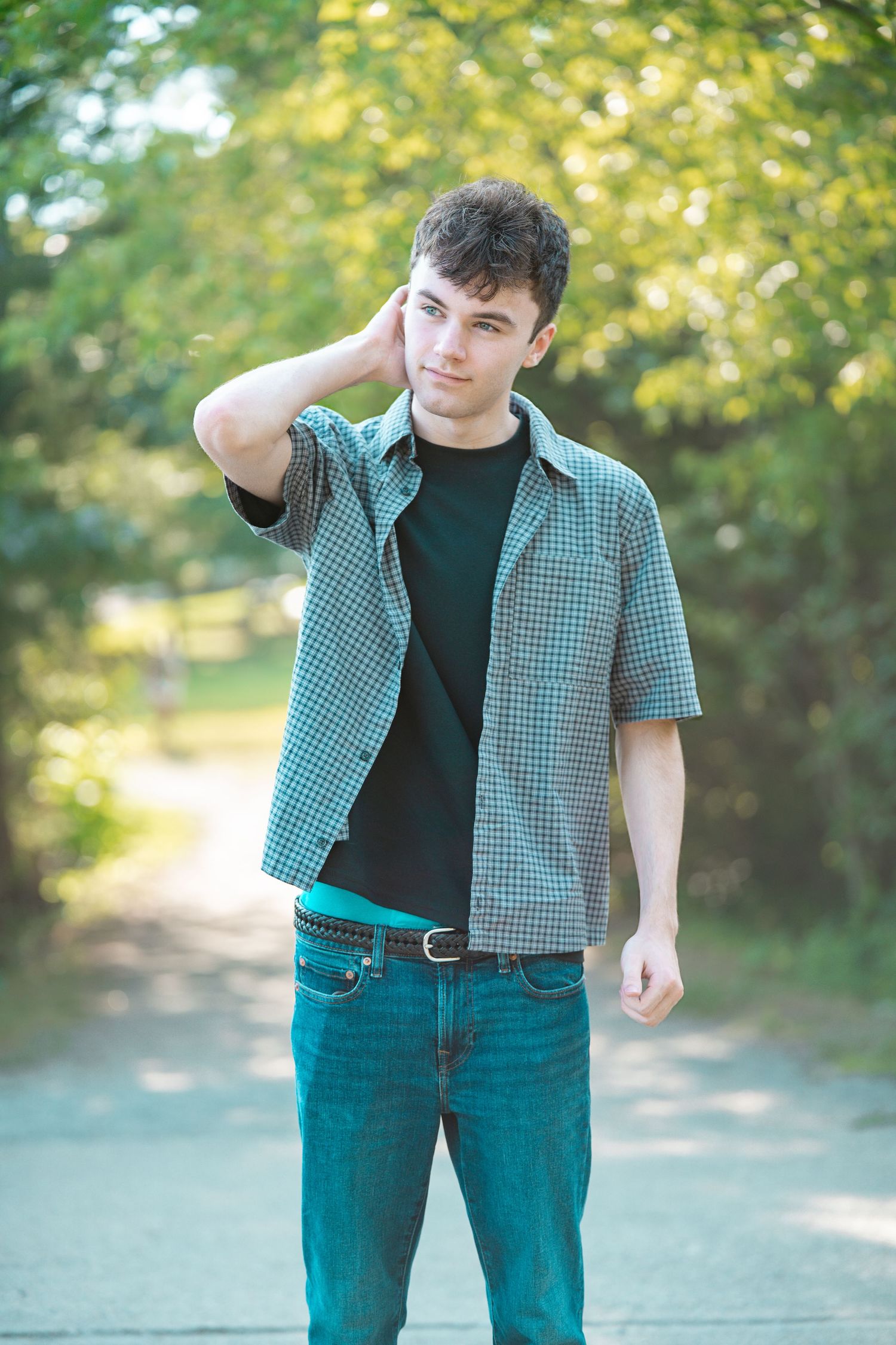 Young man in checkered shirt and jeans posing on a sunny tree-lined path.