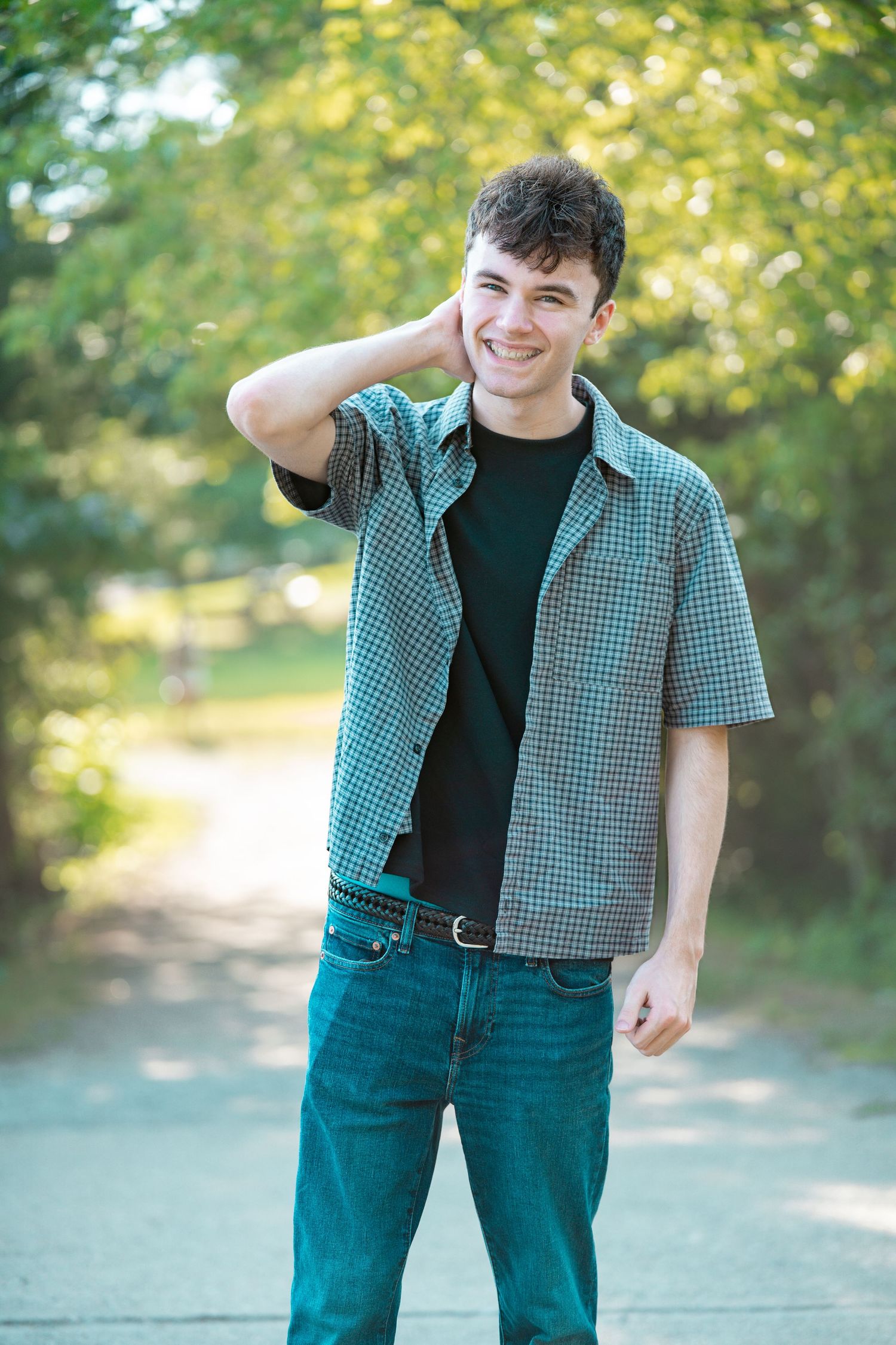 Smiling young man in a green checkered shirt and jeans stands on a sunlit tree-lined path with one hand behind his head.