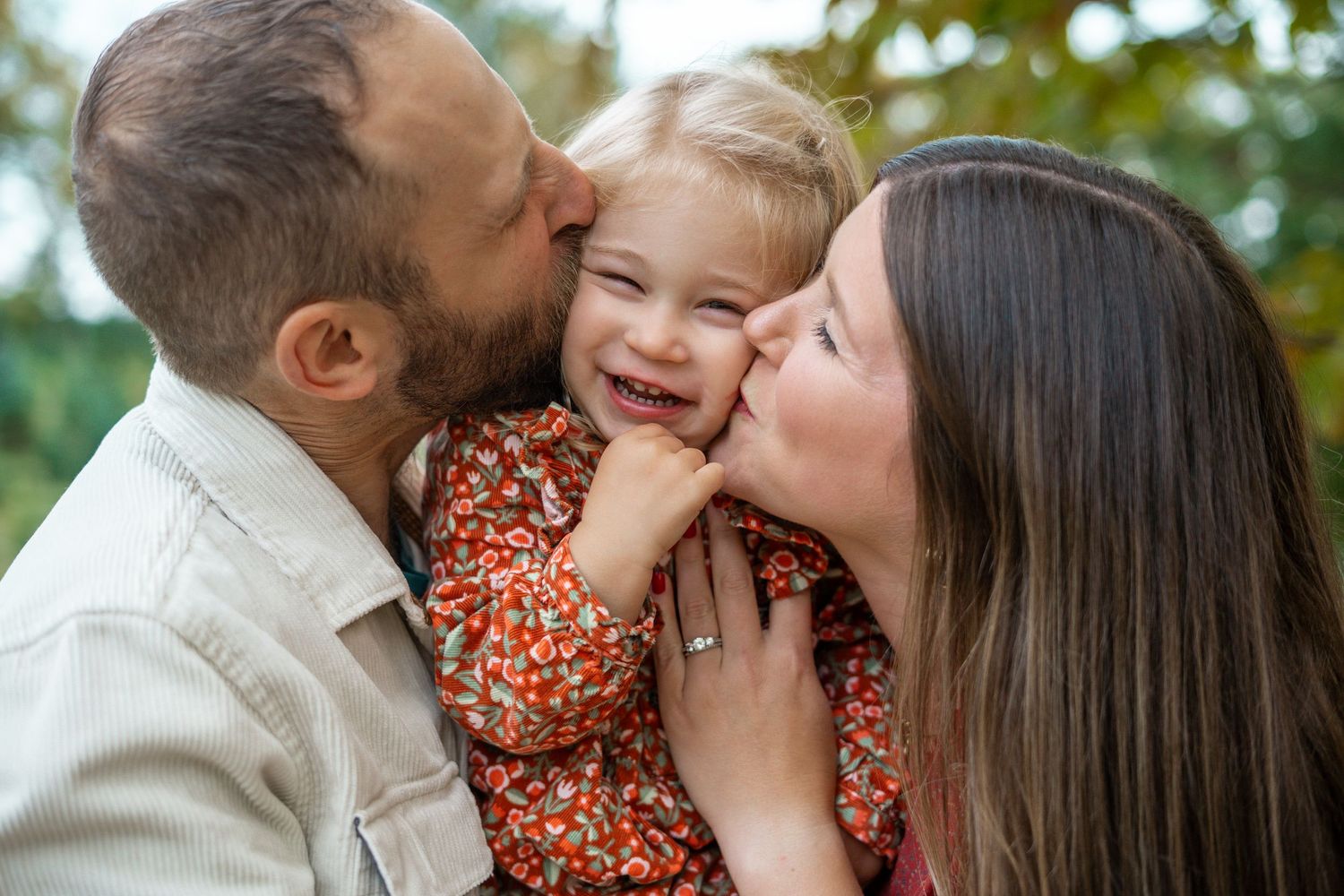 Smiling toddler in a floral top being kissed on both cheeks by her father and mother in a warm family moment outdoors.