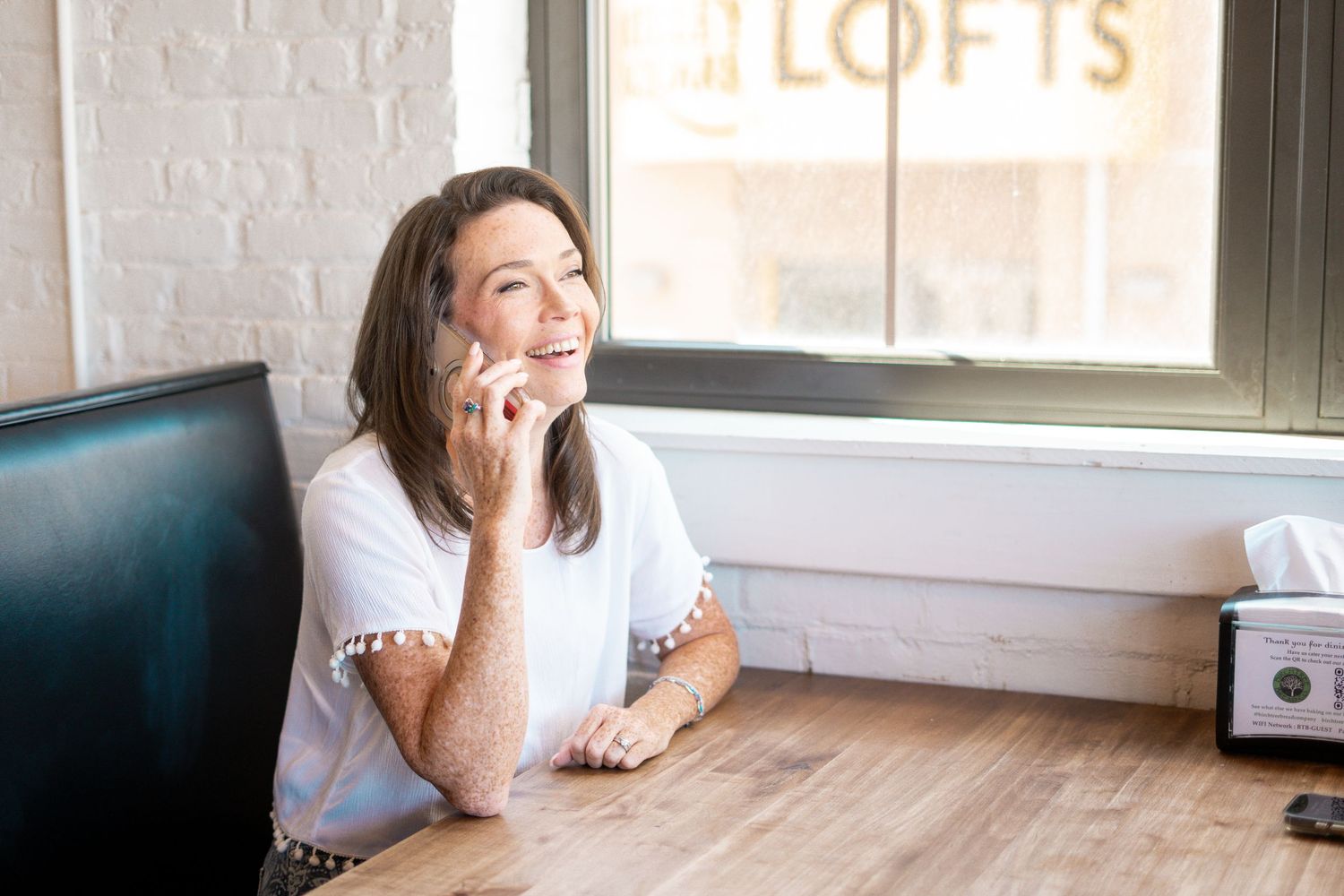 Woman smiling while talking on phone at wooden desk near window with lofts sign in bright modern space.