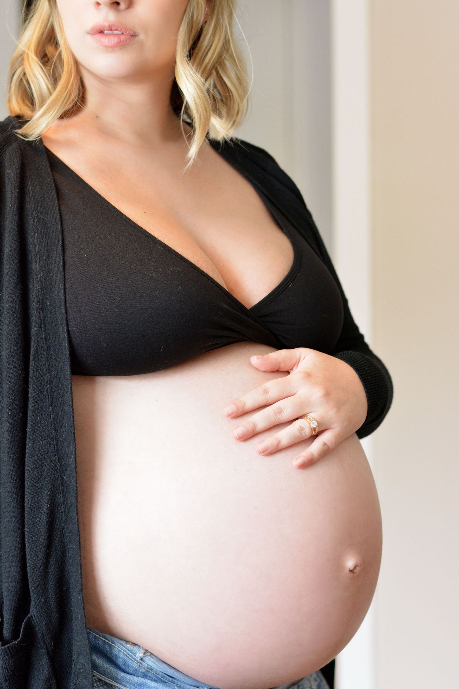 Pregnant woman in black bralette and jeans cradling baby bump indoors against neutral background.