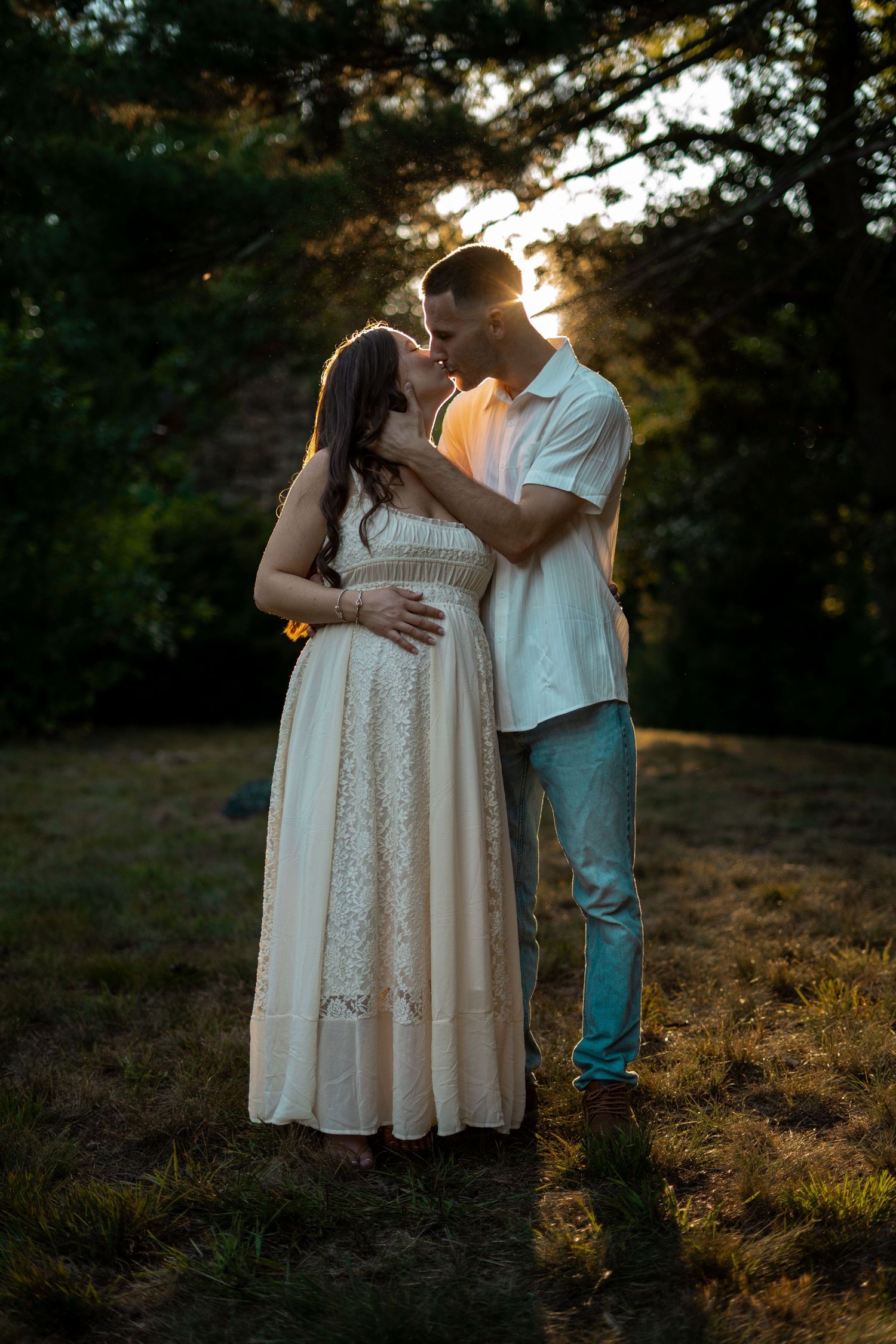 Pregnant couple sharing a kiss at golden hour sunset in a peaceful outdoor wooded setting.
