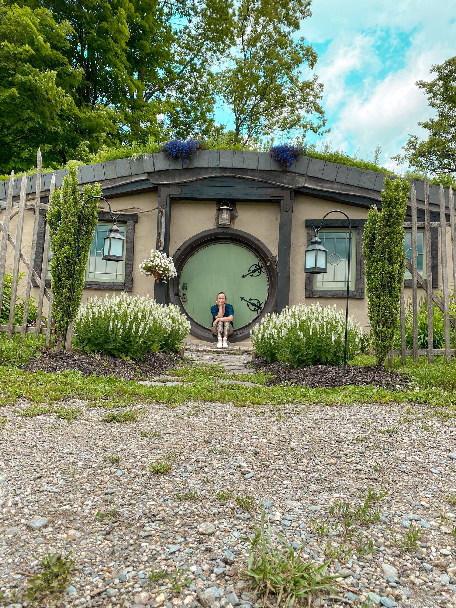 Person sitting in the doorway of a hobbit hole style building surrounded by lush greenery.