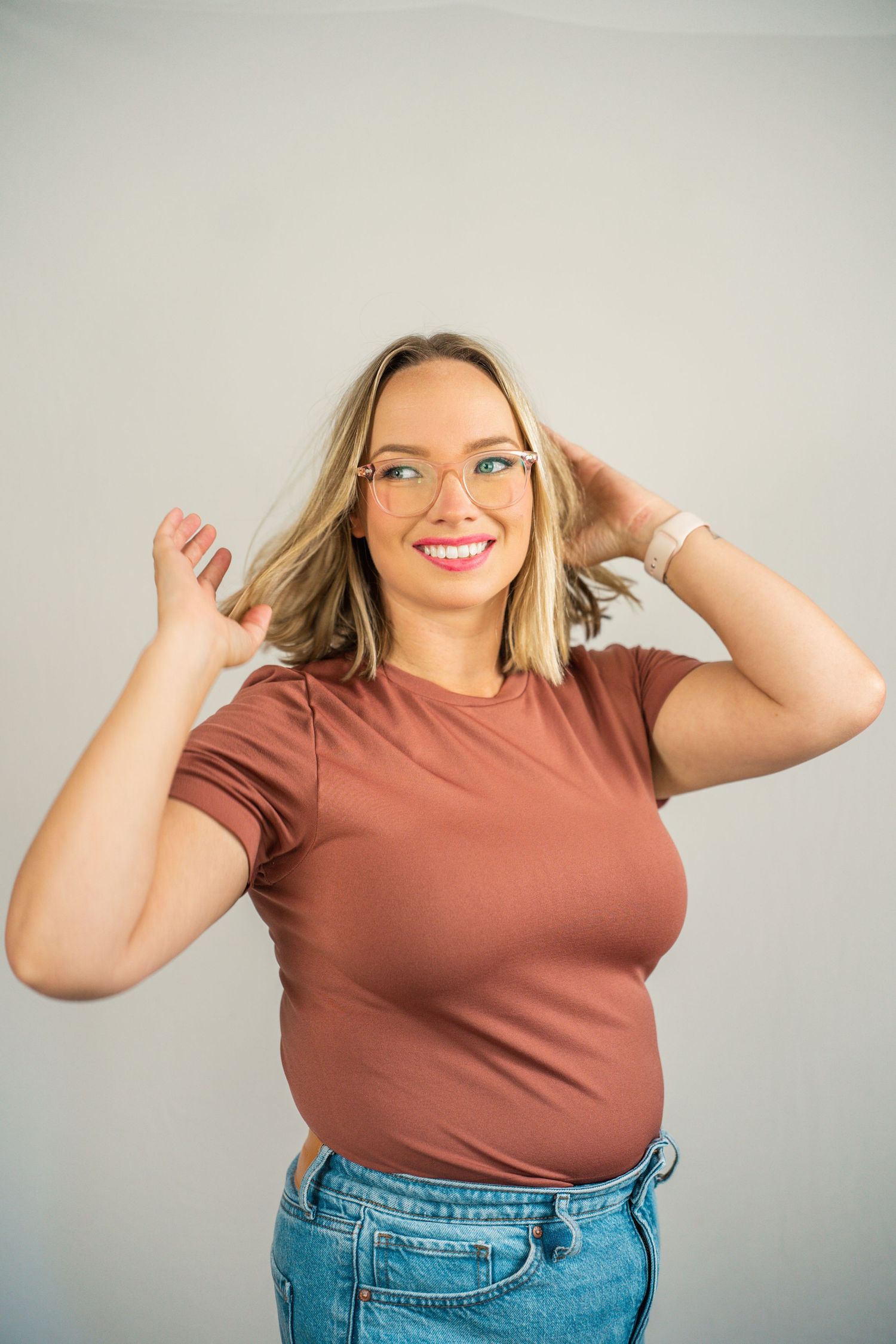 Smiling blonde woman with glasses wearing a brown t-shirt and jeans posing against a light gray background.