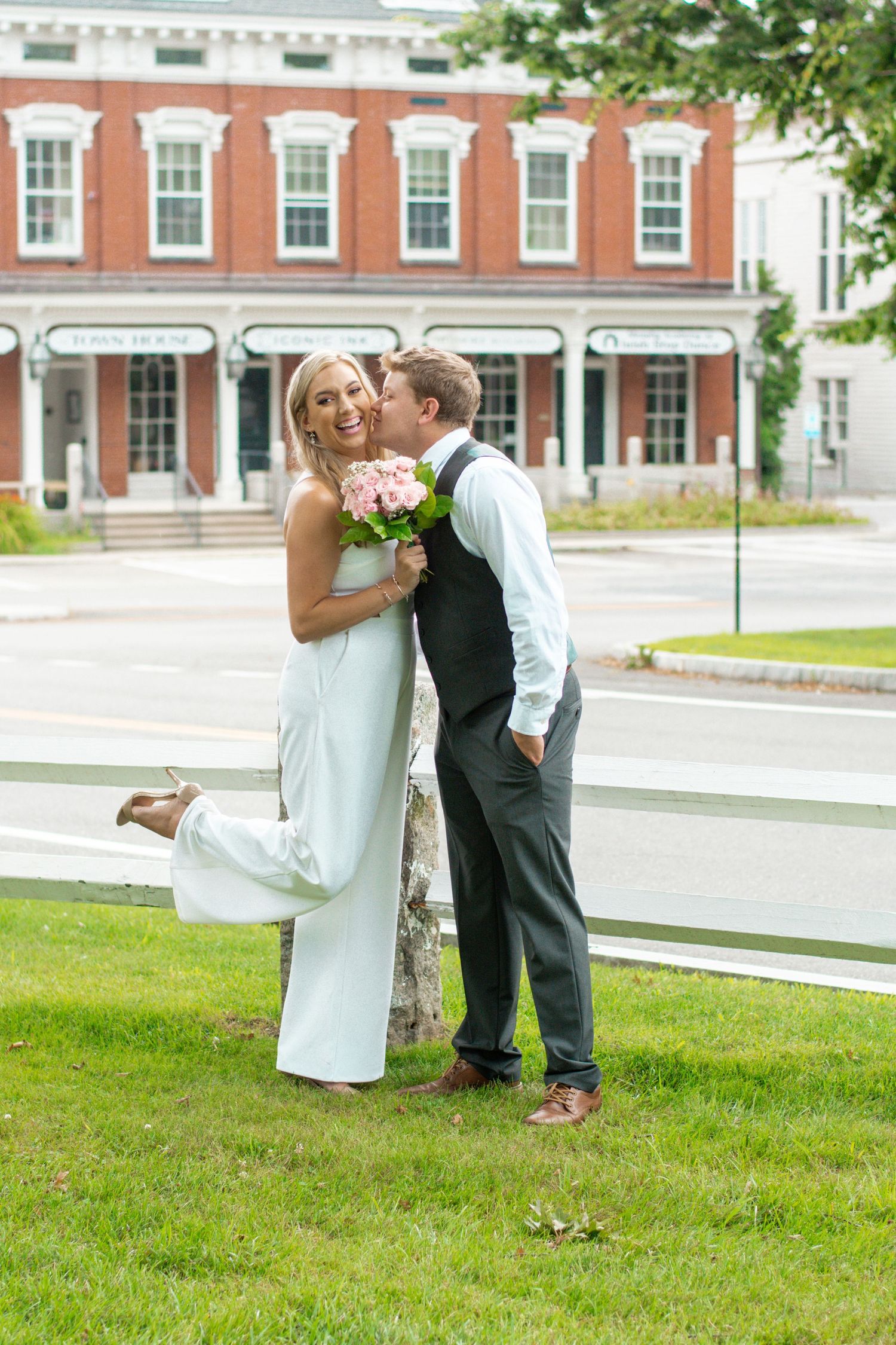 Groom kissing bride on the cheek as she laughs holding pink roses on a town green.