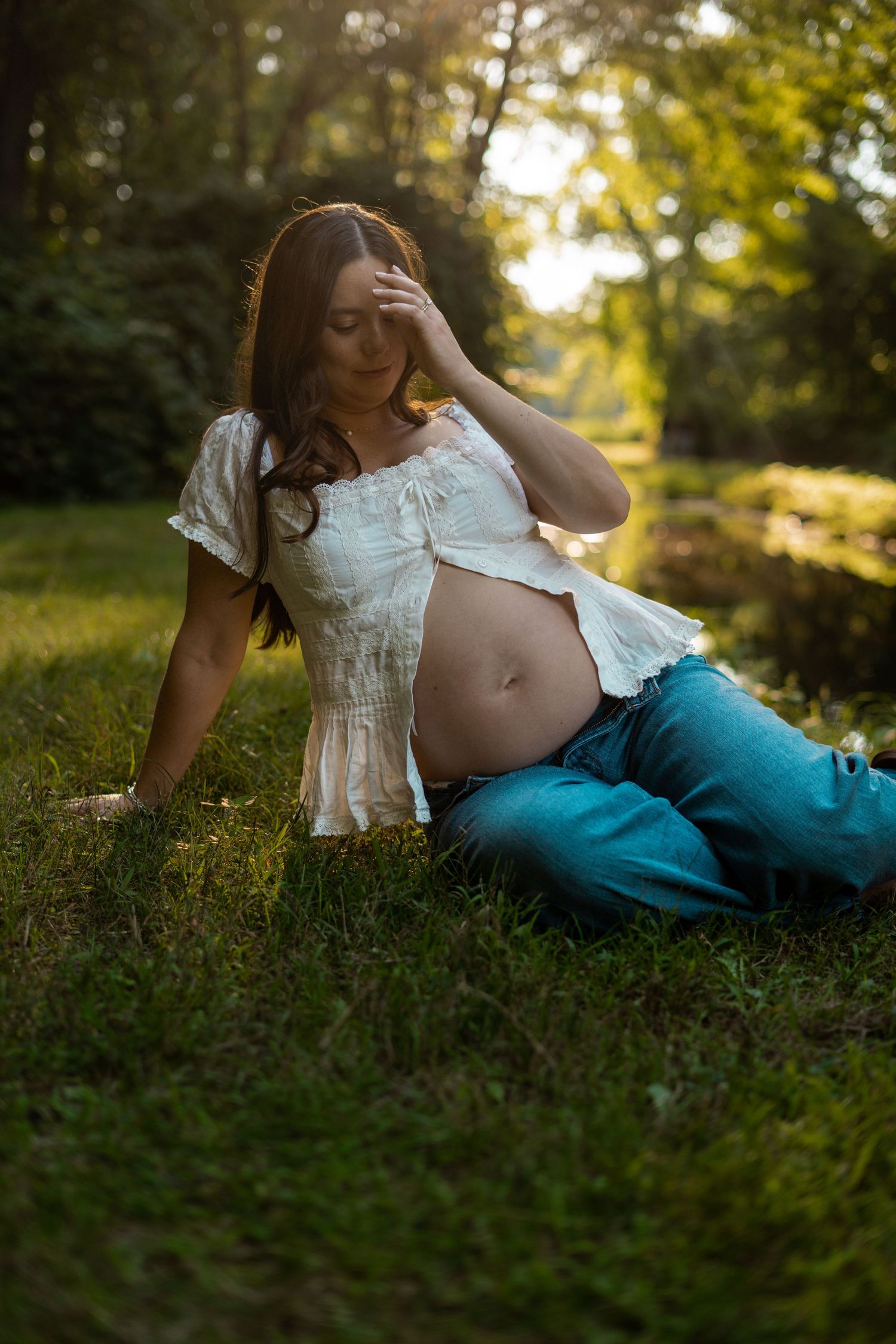 Pregnant woman in white top and jeans sitting on grass in golden sunset light outdoors.
