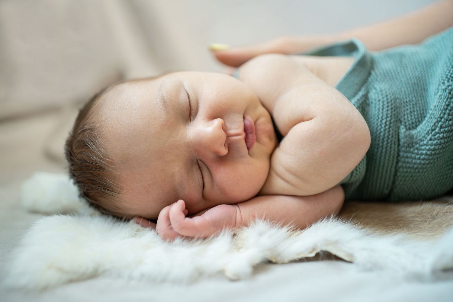 A peaceful newborn baby sleeps soundly on a white fluffy blanket wrapped in a soft teal knit wrap.