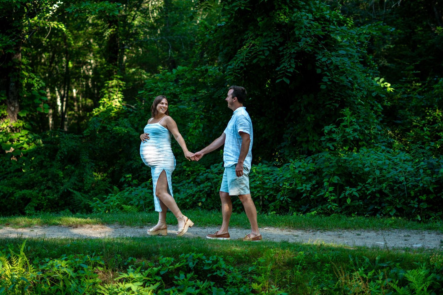 Pregnant couple holding hands and walking along a forest path during a maternity photo session.