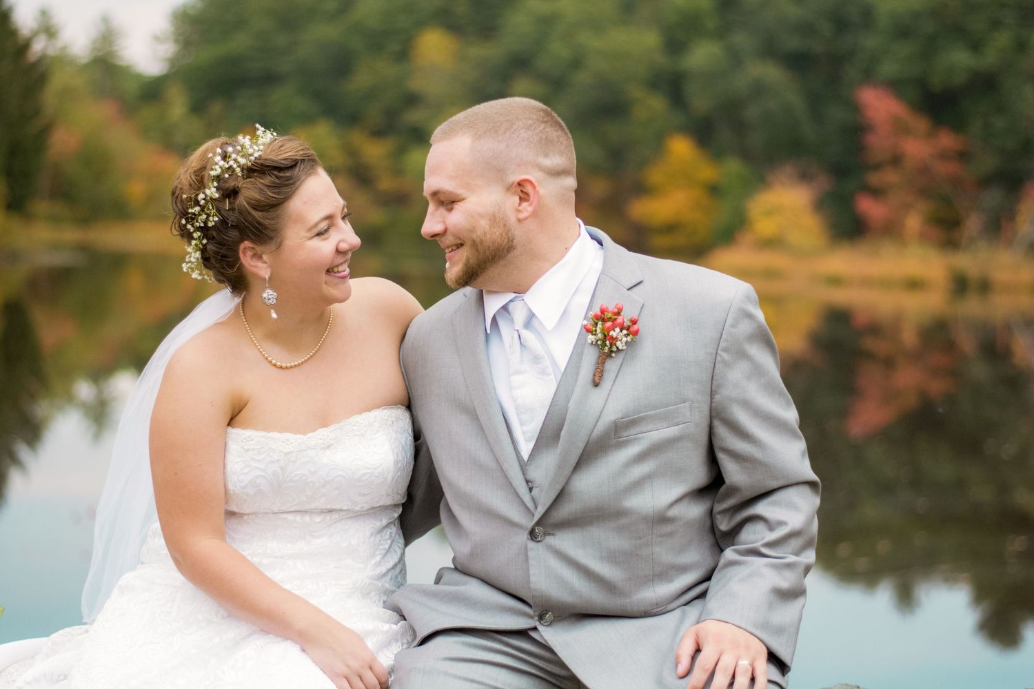 A bride and groom look at eachother sitting in front of a lake with fall foliage during this New Hampshire wedding.