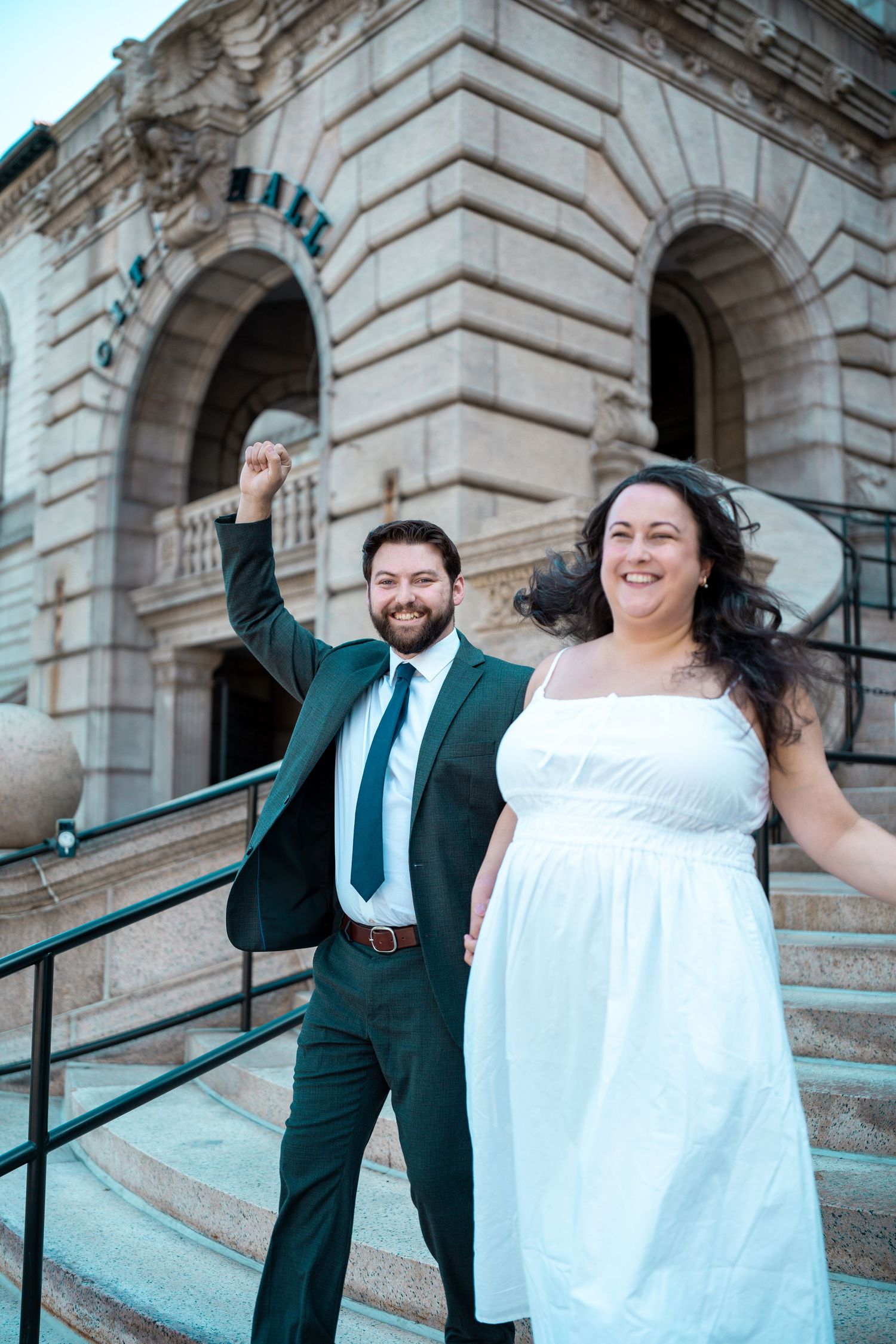 Joyful newlyweds celebrating on the steps of a grand stone hall, groom raising fist in triumph.