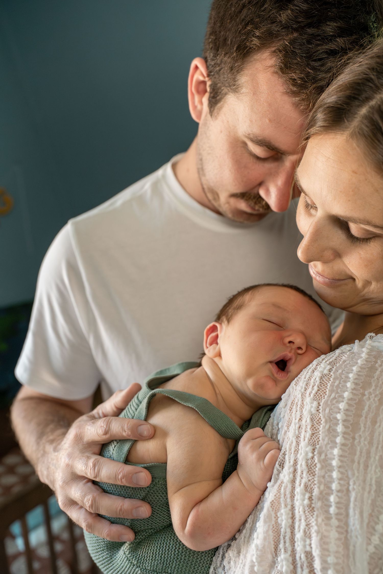 Loving parents cradling their yawning newborn baby dressed in a green outfit indoors.