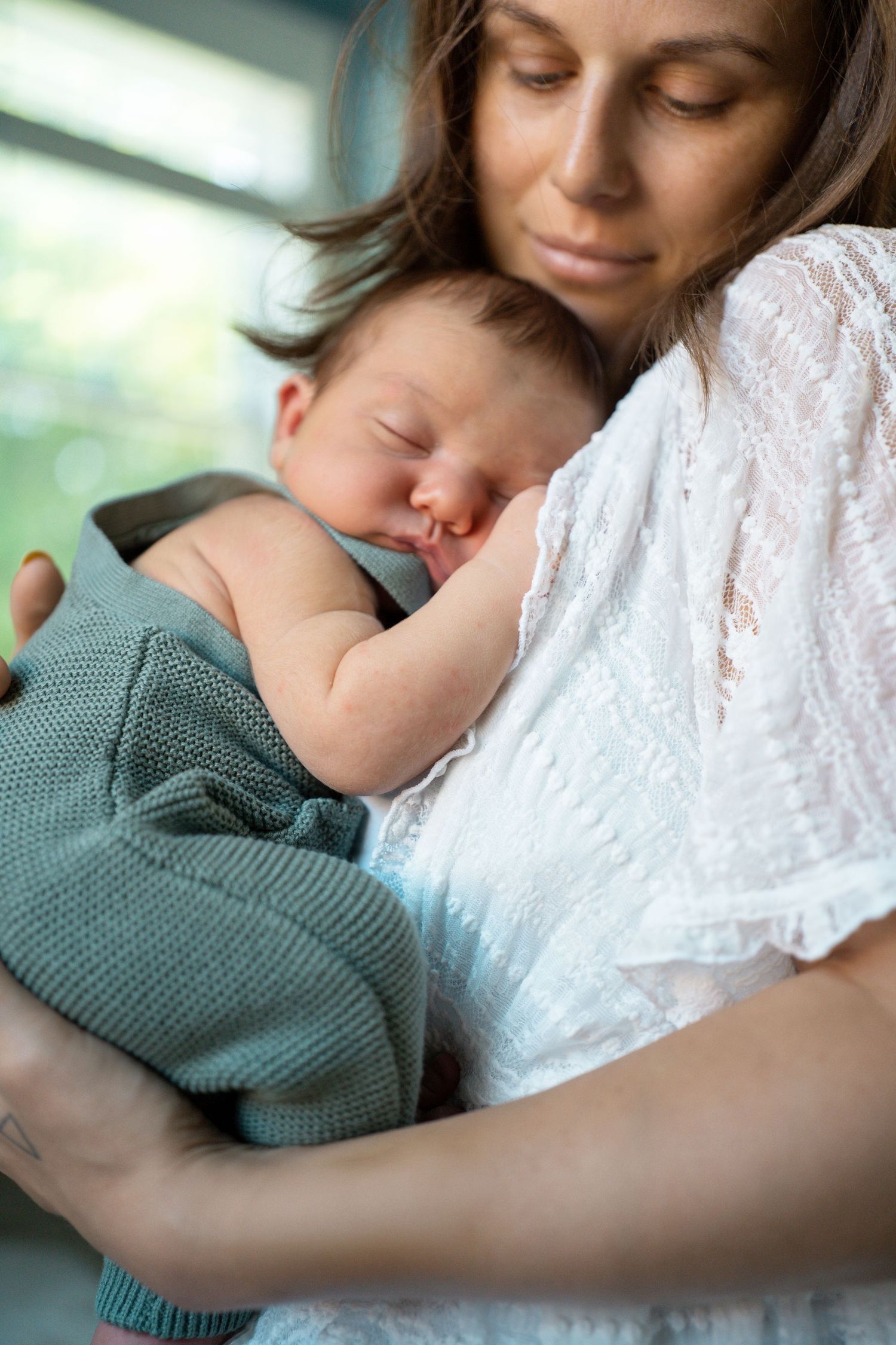 A mother gently cradles her sleeping newborn baby wrapped in a teal knit blanket near a window.