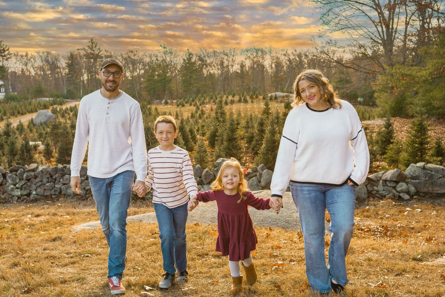 Family of four holding hands walking at a Christmas tree farm during a colorful sunset.