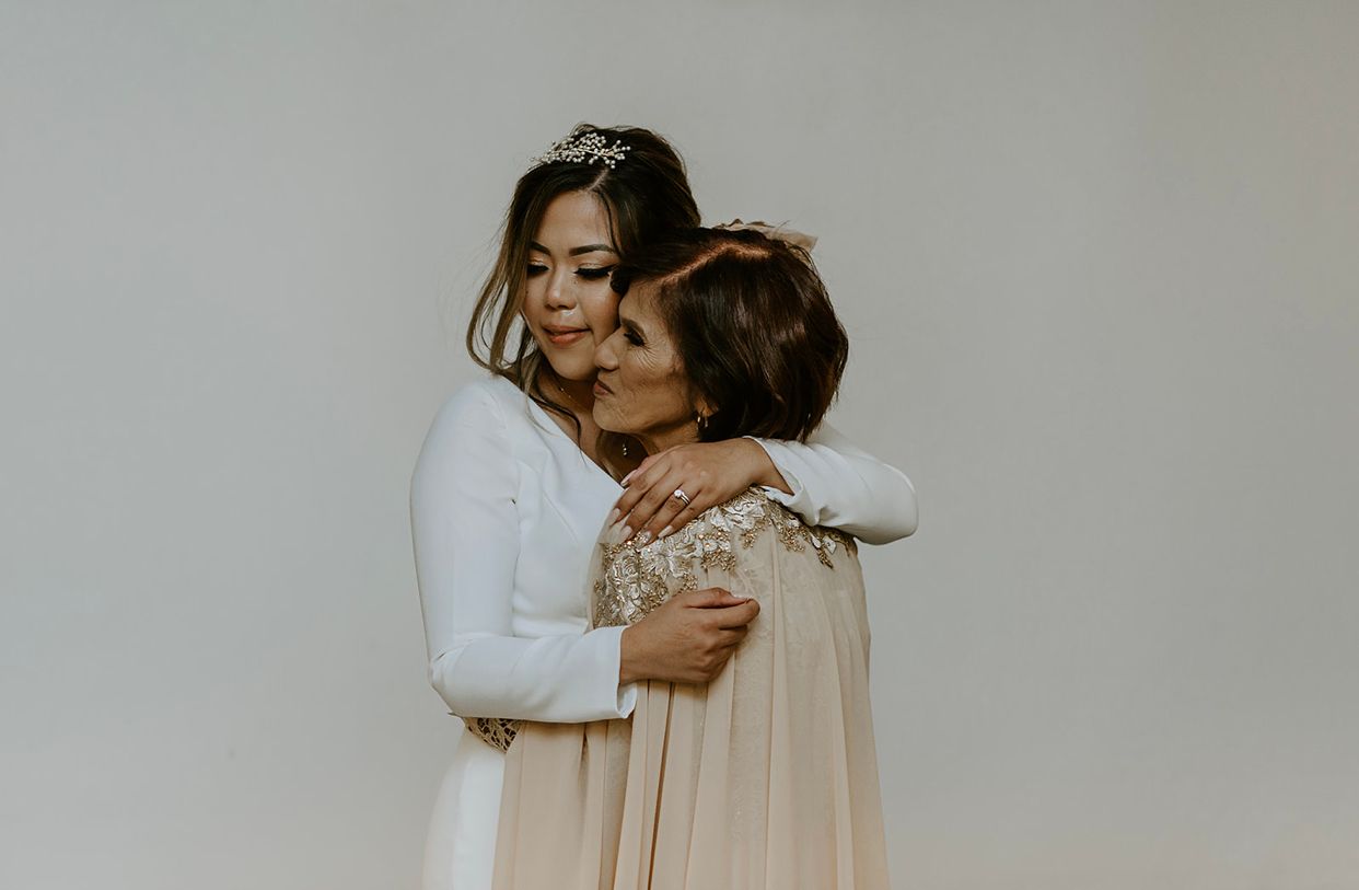 Bride in long-sleeve white gown shares tender embrace with guest in champagne dress against neutral background.
