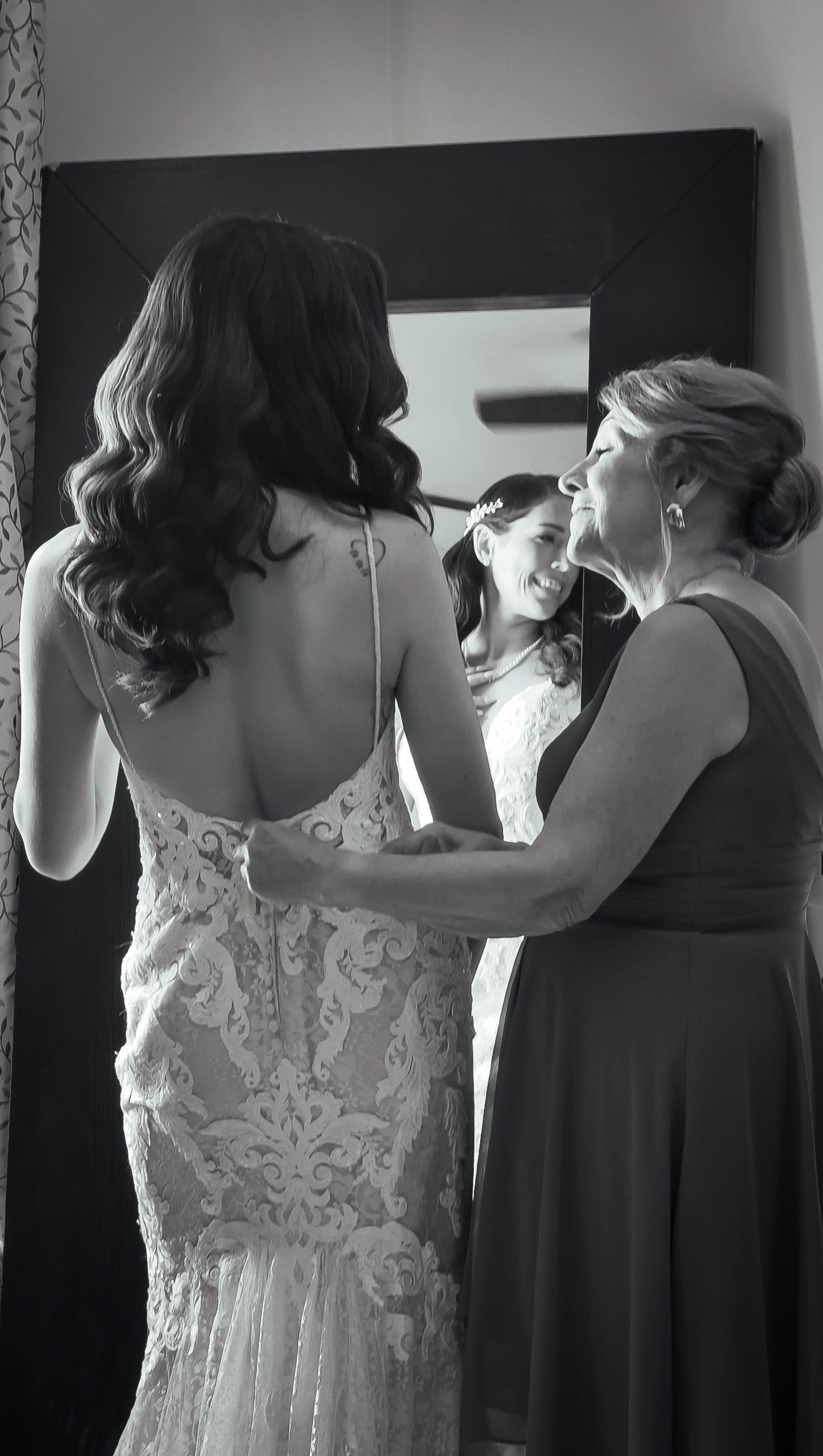 Black and white photo of a bride in a lace gown being helped by two women smiling in a mirror reflection.