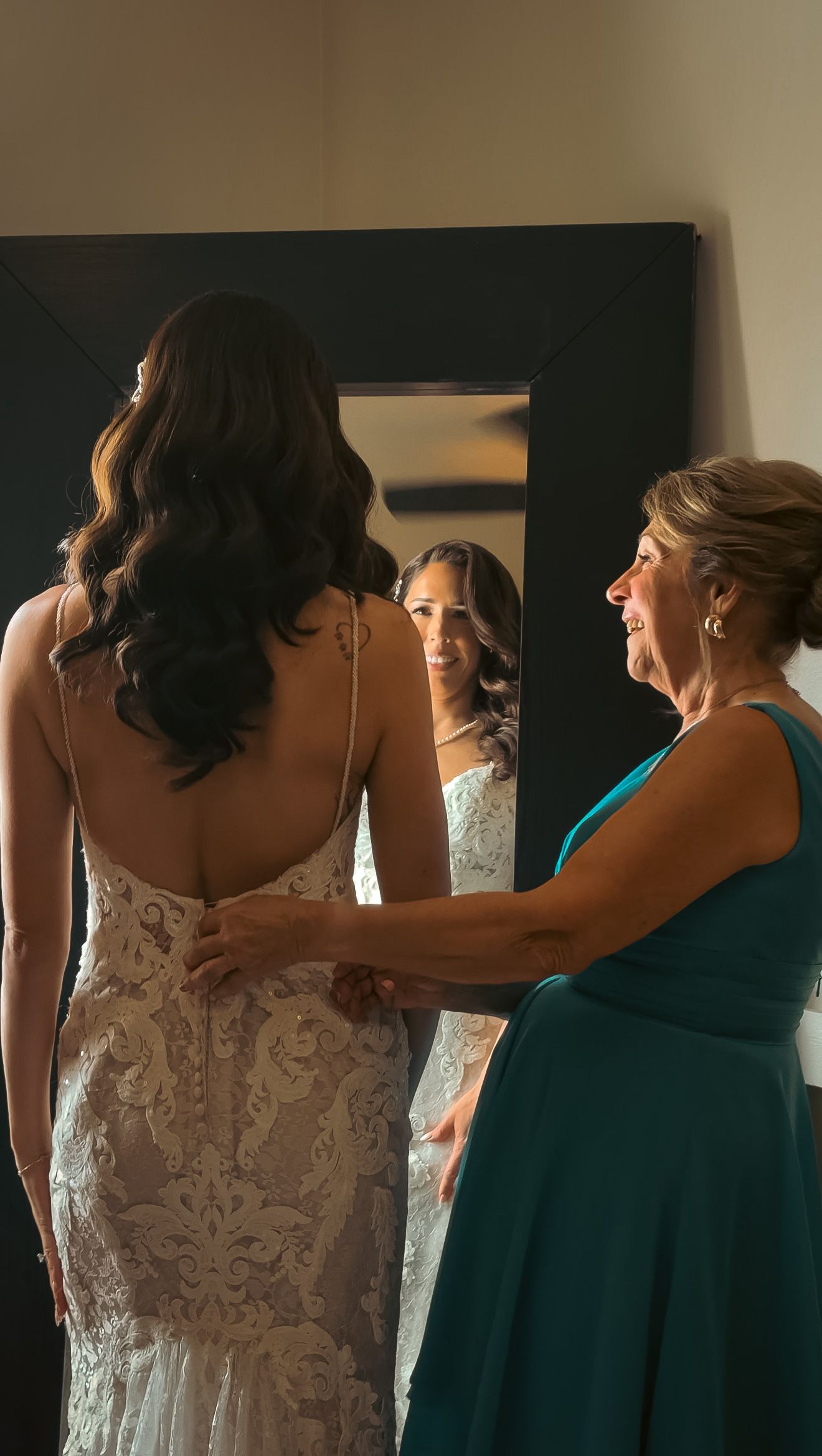 Mother in teal dress helping bride in lace backless gown prepare in front of mirror.