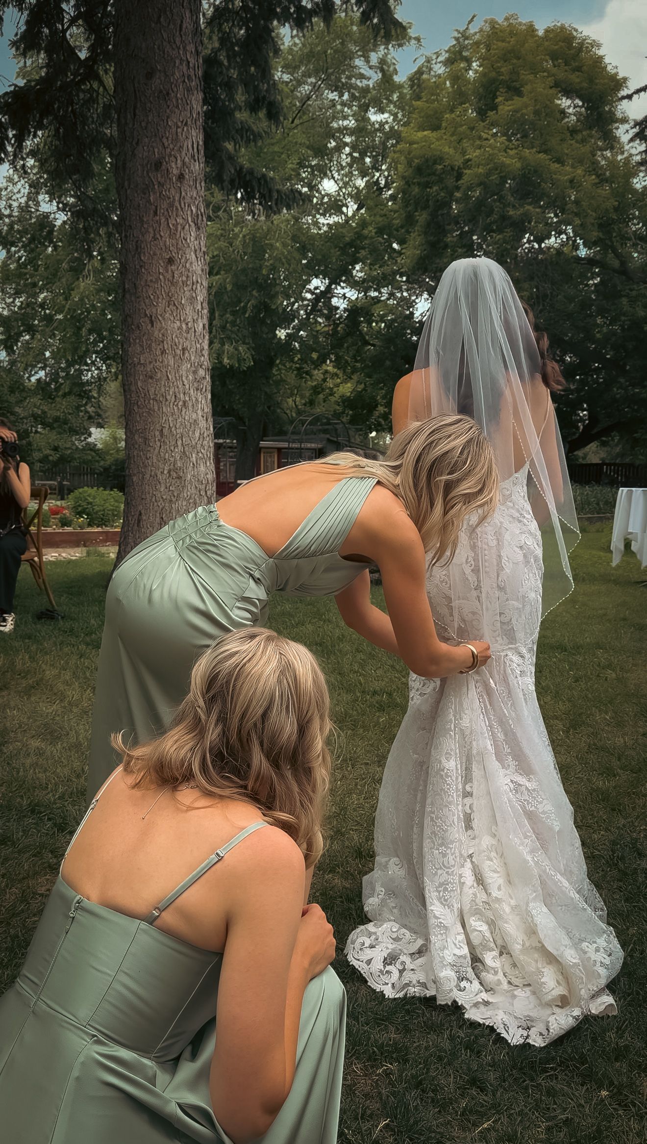 Bridesmaids in sage green gowns adjusting bride's lace wedding dress and veil outdoors.