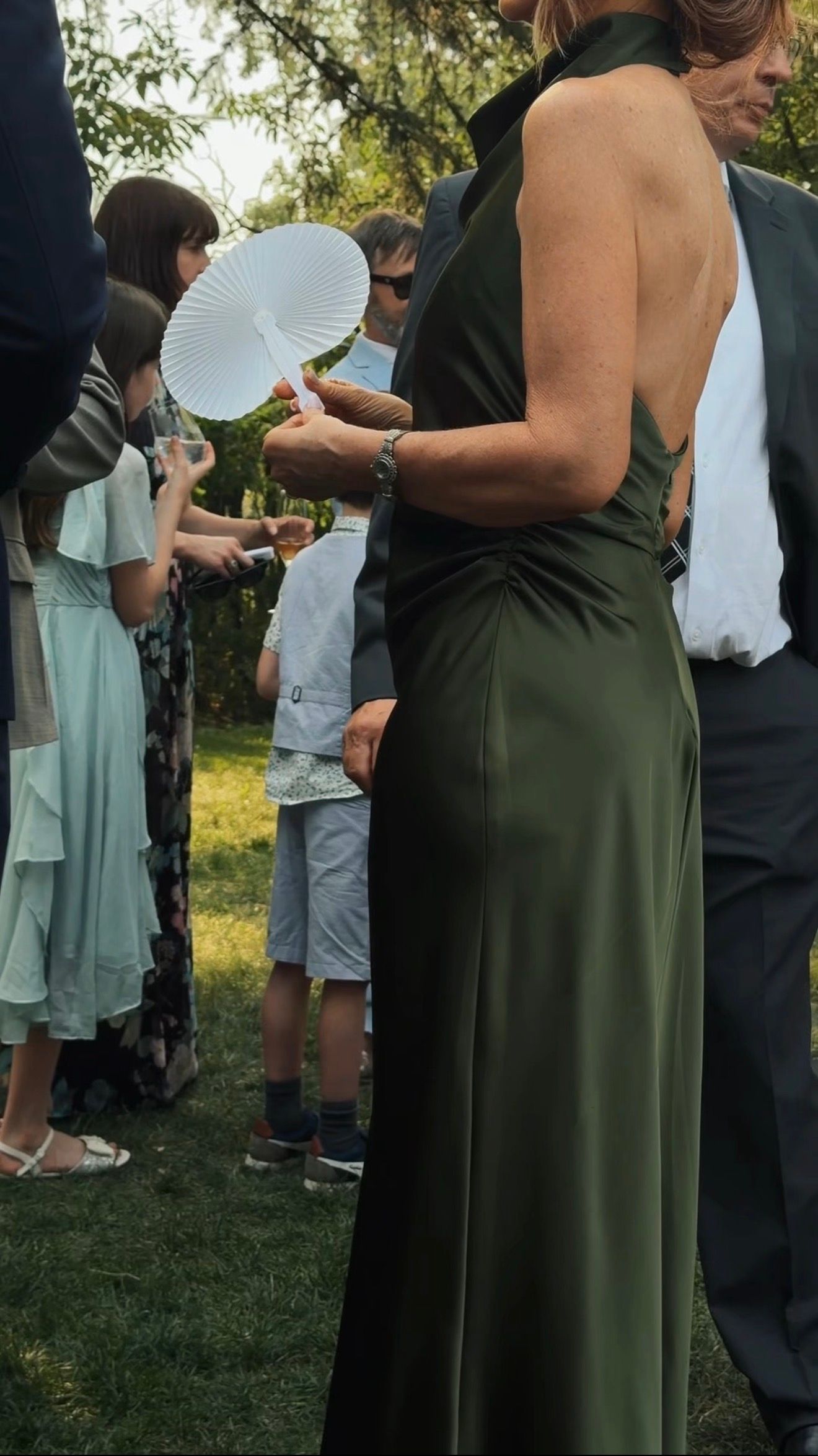 Woman in elegant backless olive green halter gown holds white paper fan at outdoor wedding reception.