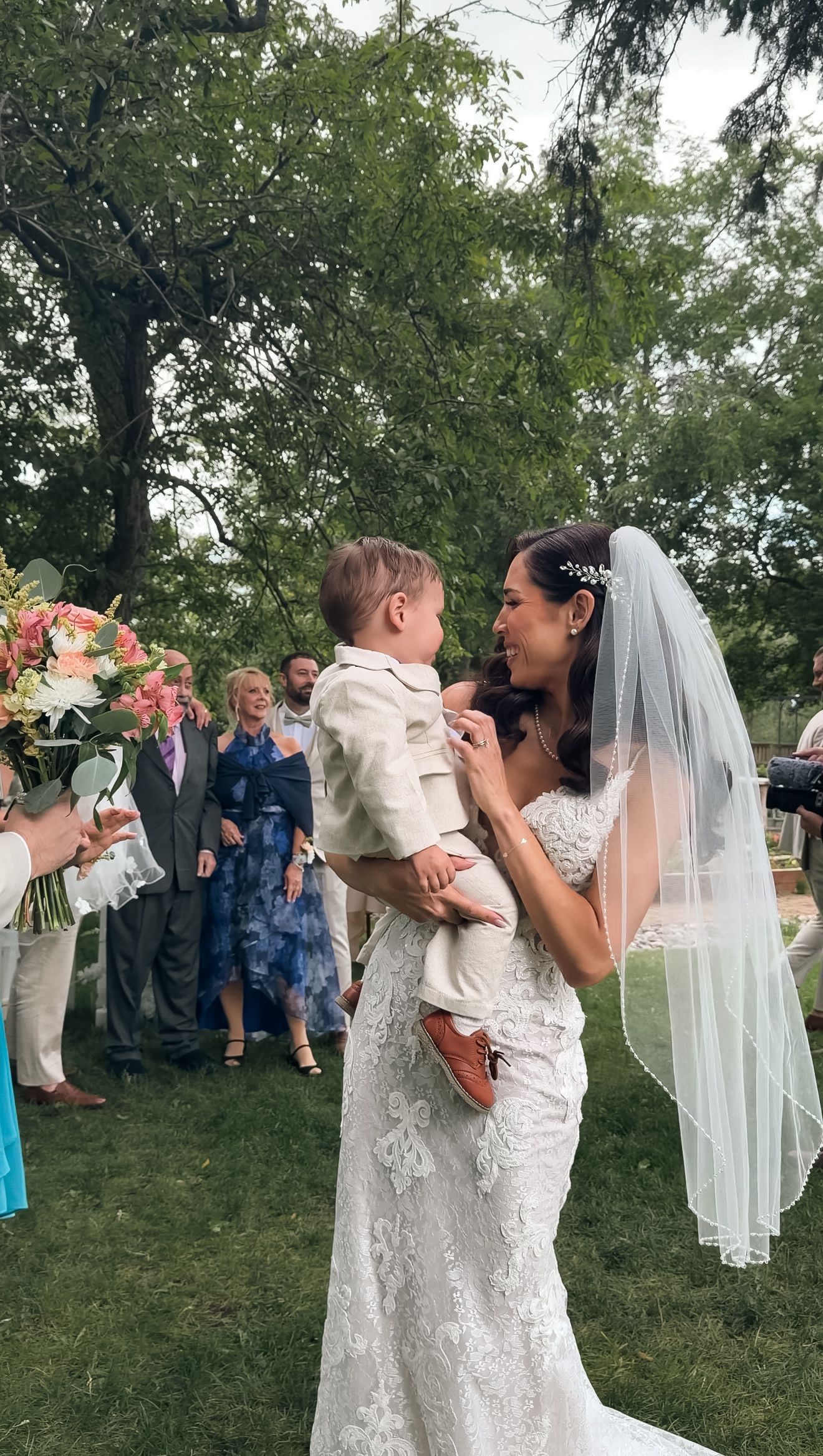 Radiant bride in lace gown and veil holds young child while guests celebrate after outdoor ceremony.