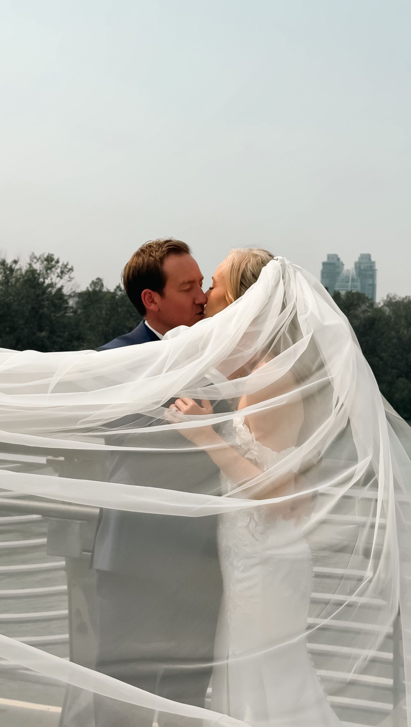 Newlyweds share romantic kiss as bride's veil billows dramatically in the wind on rooftop.