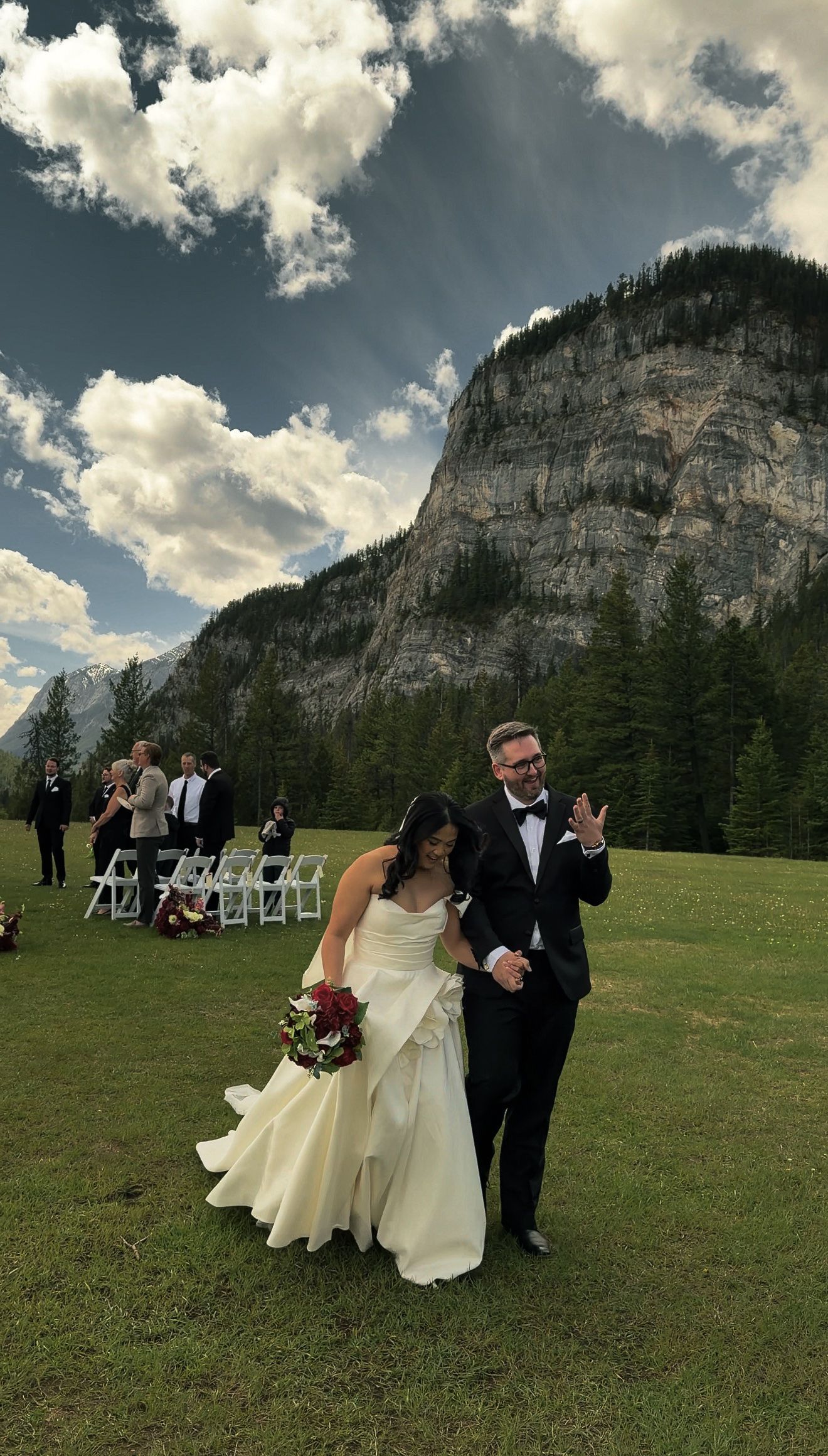 Newlywed couple walks joyfully after outdoor mountain wedding ceremony in Banff with guests applauding behind them.