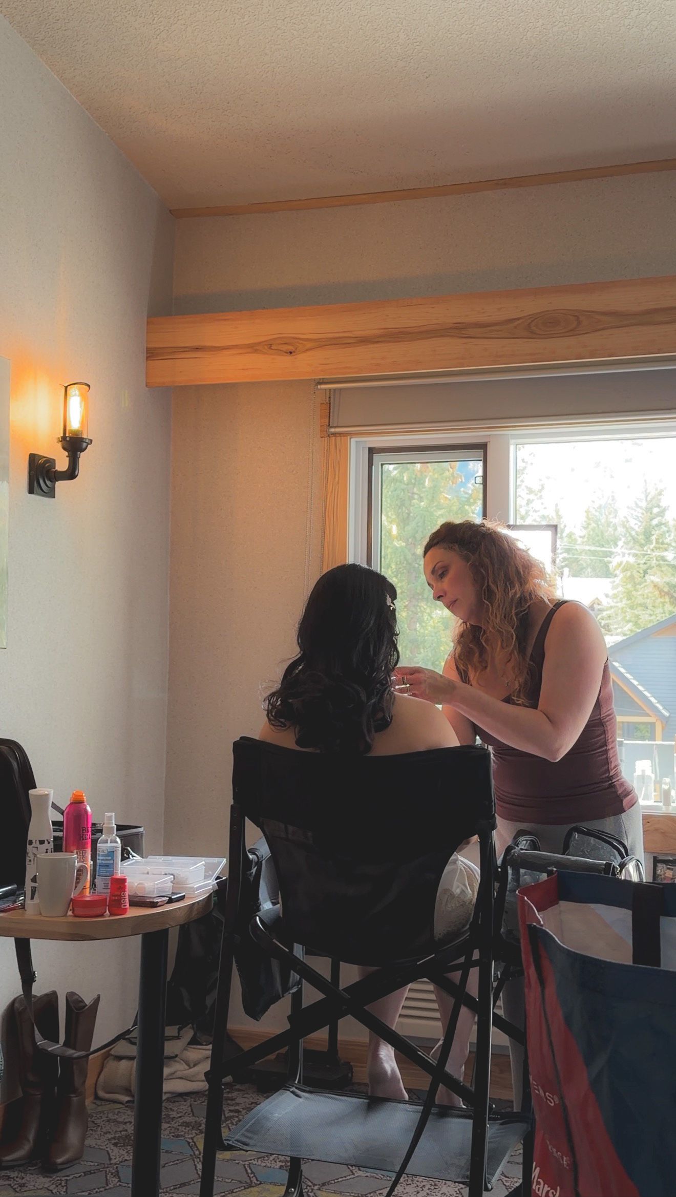 A makeup artist applies finishing touches to a dark-haired woman seated in a director's chair near a sunlit window.
