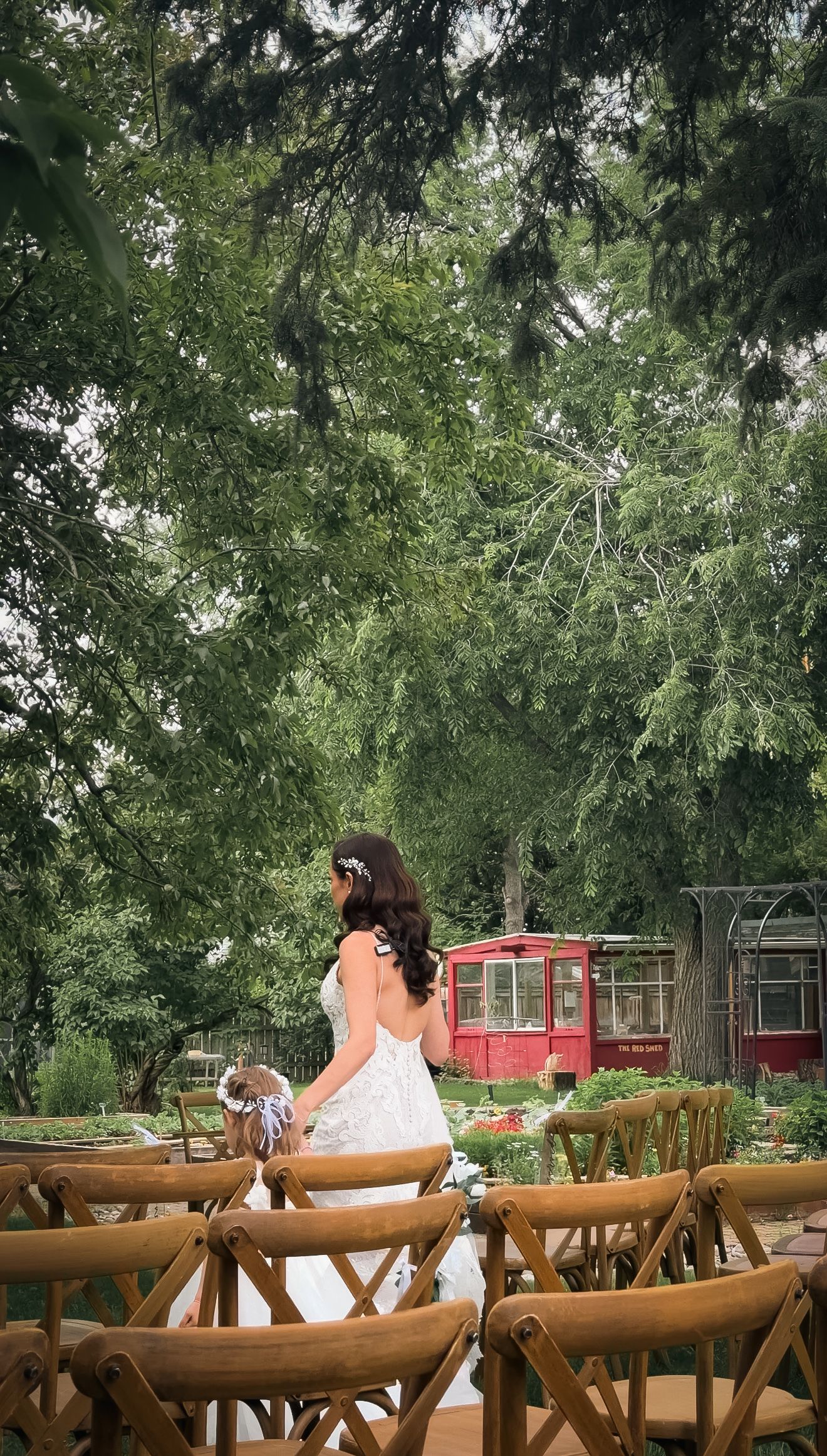 Bride in lace gown walks down aisle between wooden chairs toward lush green tree-lined ceremony space.